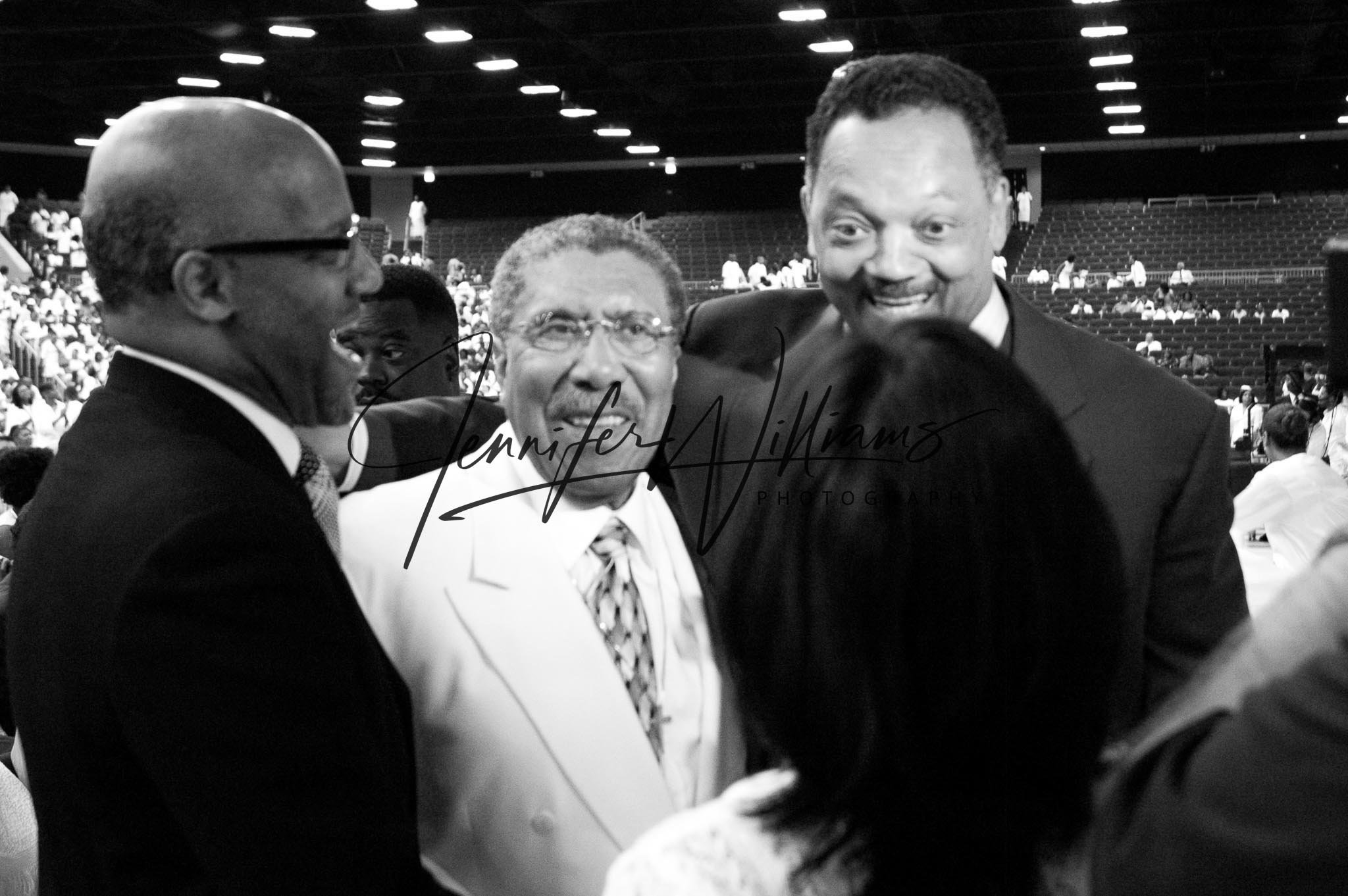 Black and white photo of four people in suits talking and smiling at each other in an indoor arena with many seated onlookers in the background.