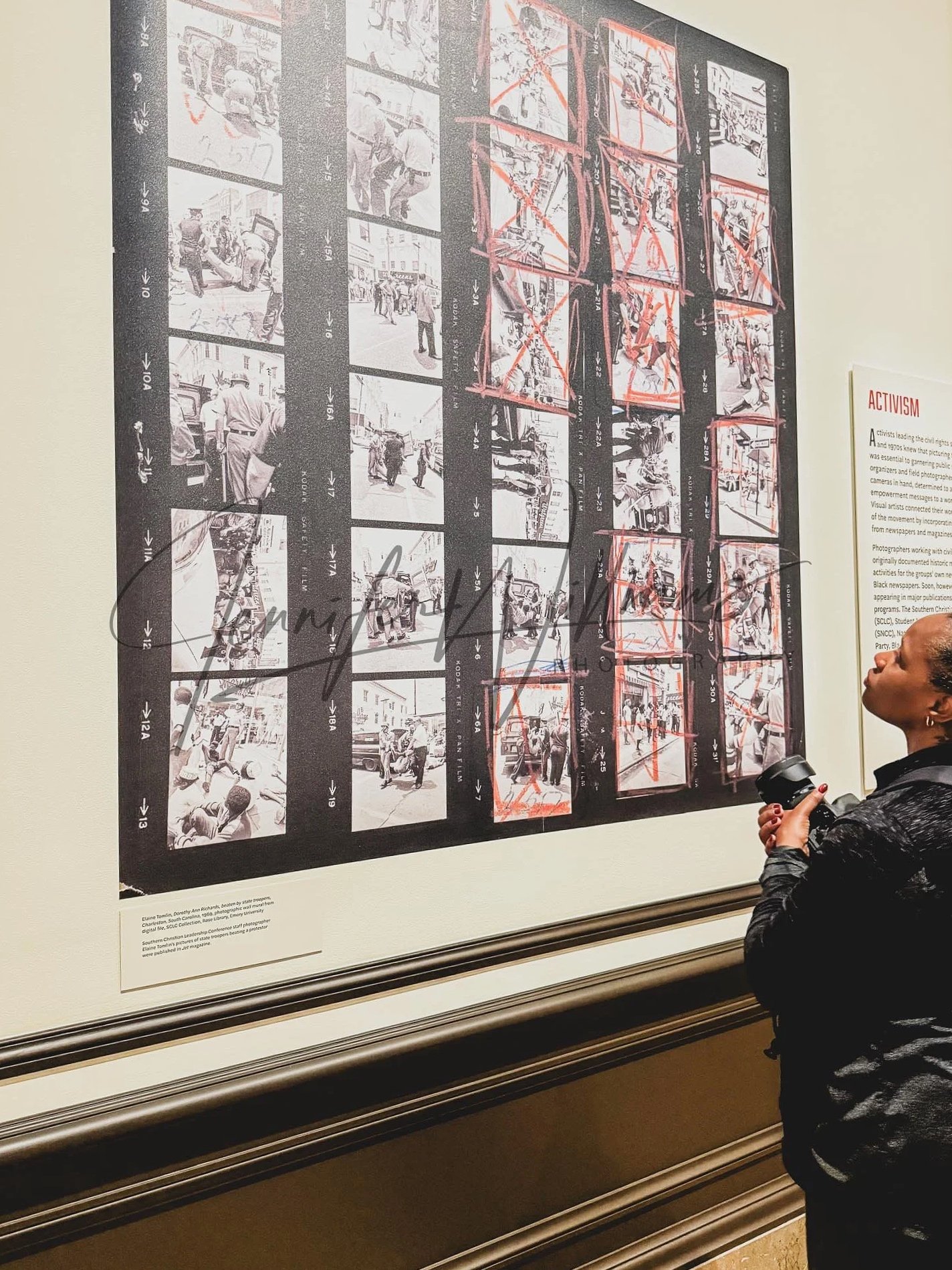 A woman stands in front of a large black-and-white photo collage on a wall, looking at it and holding a camera in her right hand. The collage contains multiple photographs, some marked with red crosses and lines, and has section labels with numbers and arrows.
