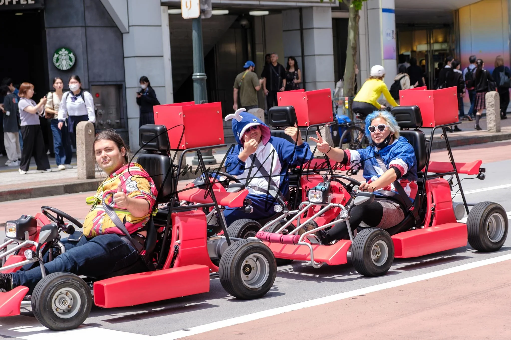Three people riding go-karts on a city street, with pedestrians walking in the background. The individuals are dressed in colorful, casual clothing, and appear to be enjoying themselves.