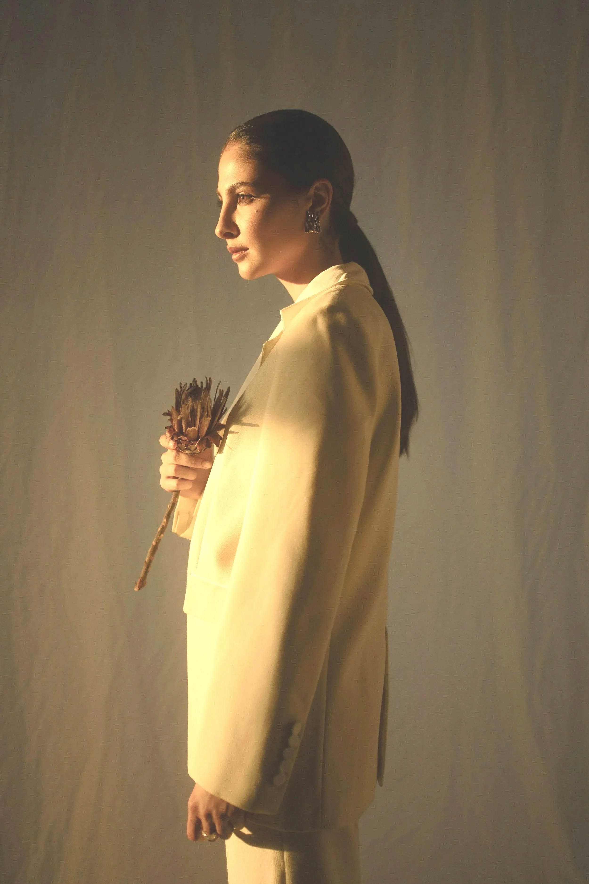 Une femme portant un tailleur beige, tenant une branche avec une fleur, regardant sur le côté, éclairée par une lumière douce dans un fond neutre.