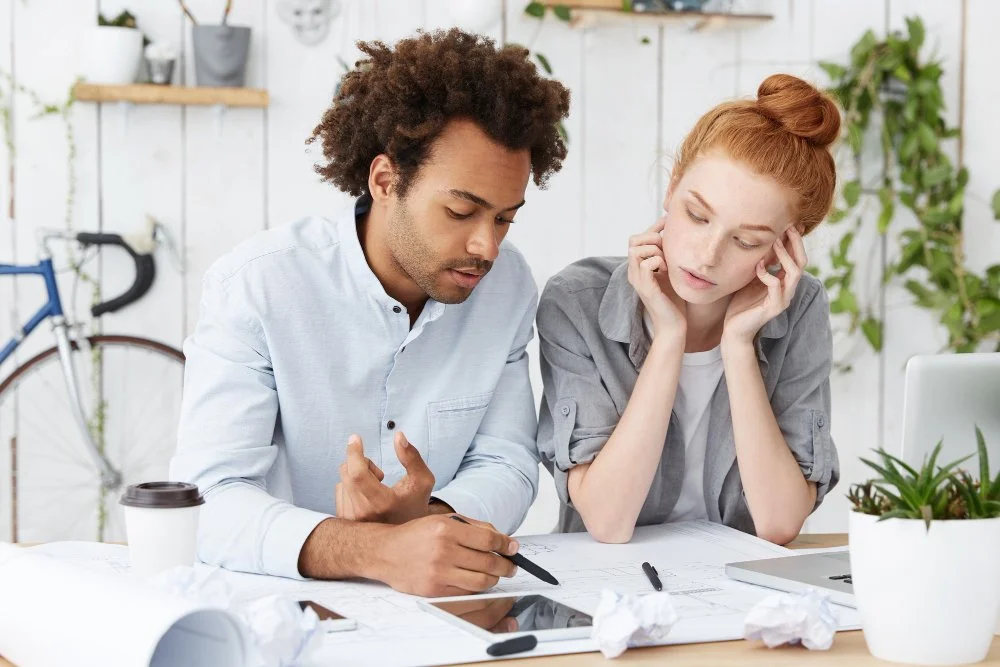 Two people, a man with curly hair and a woman with red hair, are sitting at a desk looking at a tablet and discussing. The desk has papers, a pen, crumpled papers, and a coffee cup. In the background, there's a bicycle and green plants.