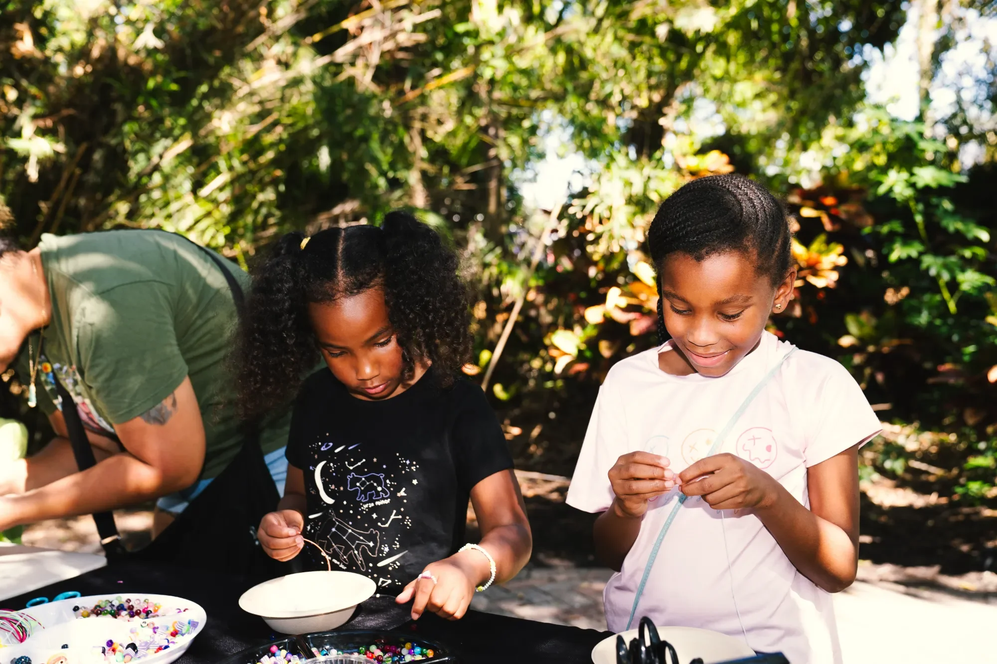 Two young girls and an adult woman participate in a bead craft activity outdoors among trees. The girls are focused on stringing beads while the woman assists. They are surrounded by bowls of colorful beads and craft supplies.