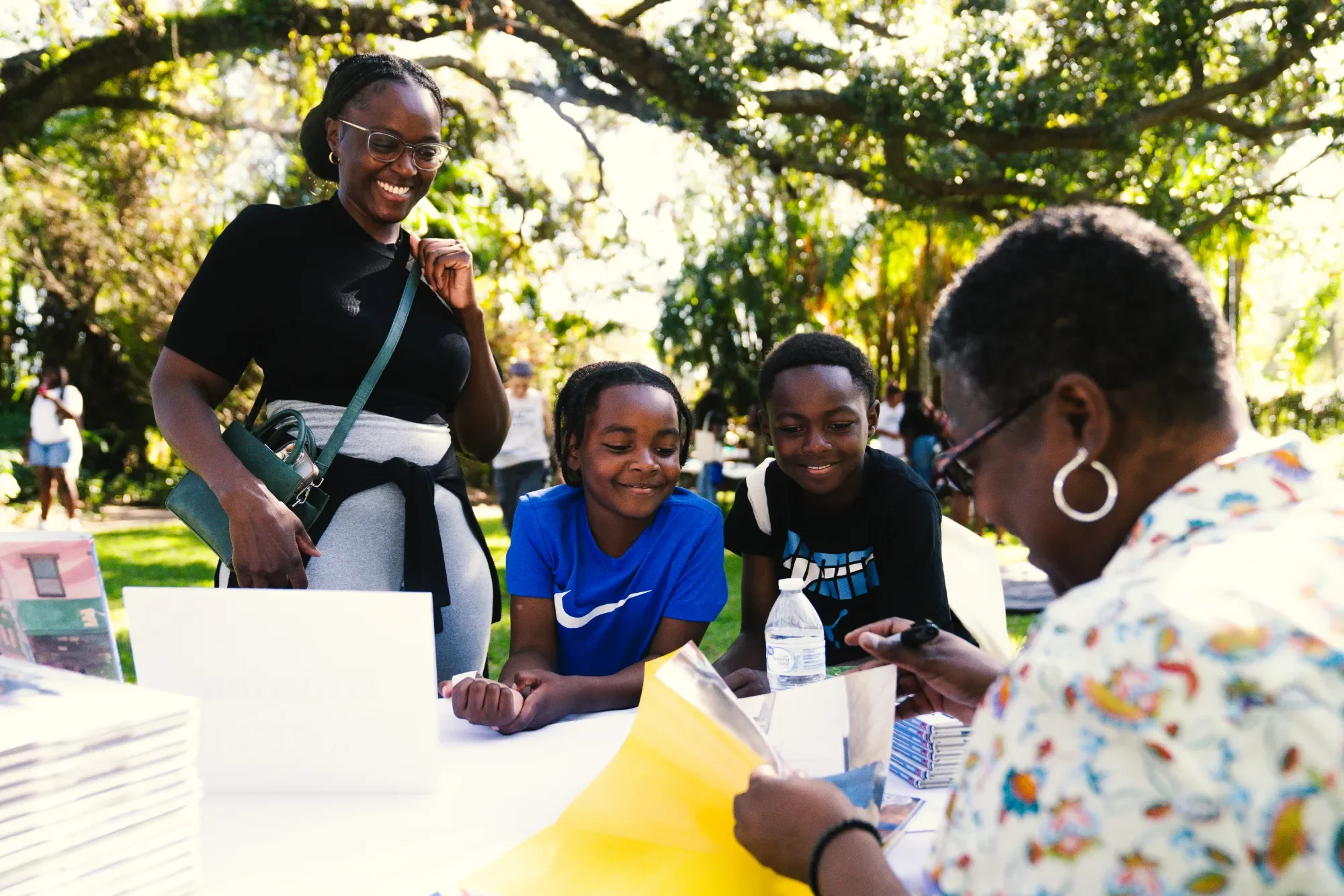 Group of people at an outdoor event, with a woman smiling and standing next to children who are looking at a phone and books on a table. There are trees in the background and sunlight filtering through the leaves.