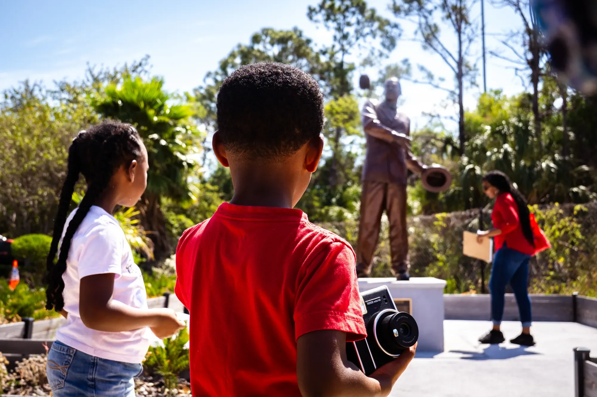 Children attending an outdoor event, with one boy in a red shirt holding a camera, and girls nearby, listening to a man with a hat playing a guitar, in a lush garden setting on a sunny day.
