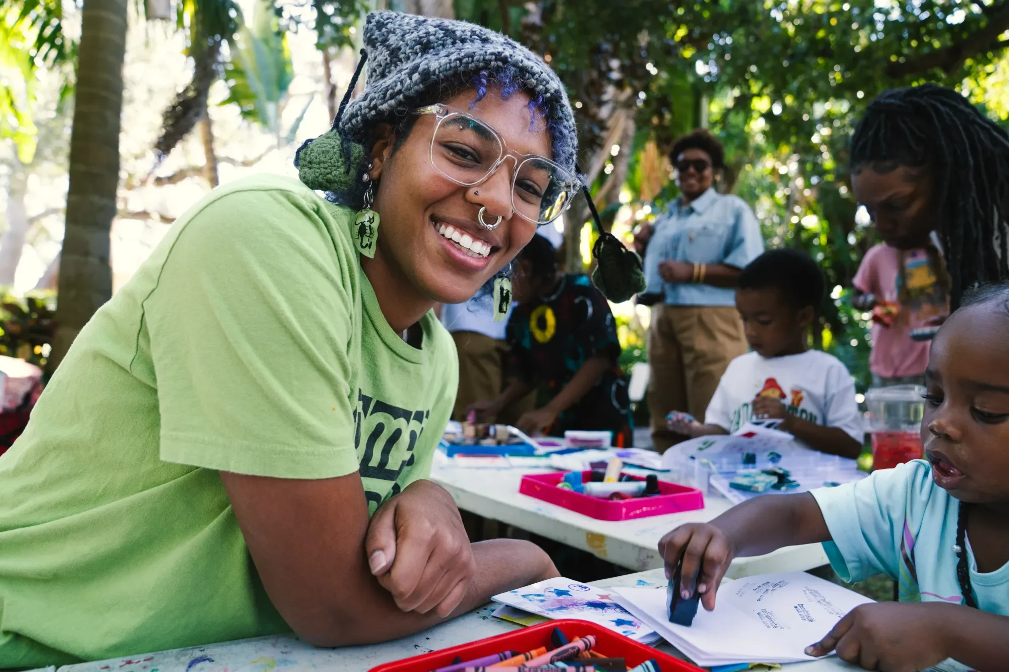 A woman with blue and purple hair, glasses, and a nose ring, smiling at a group of children at an outdoor arts and crafts activity, with trees in the background.
