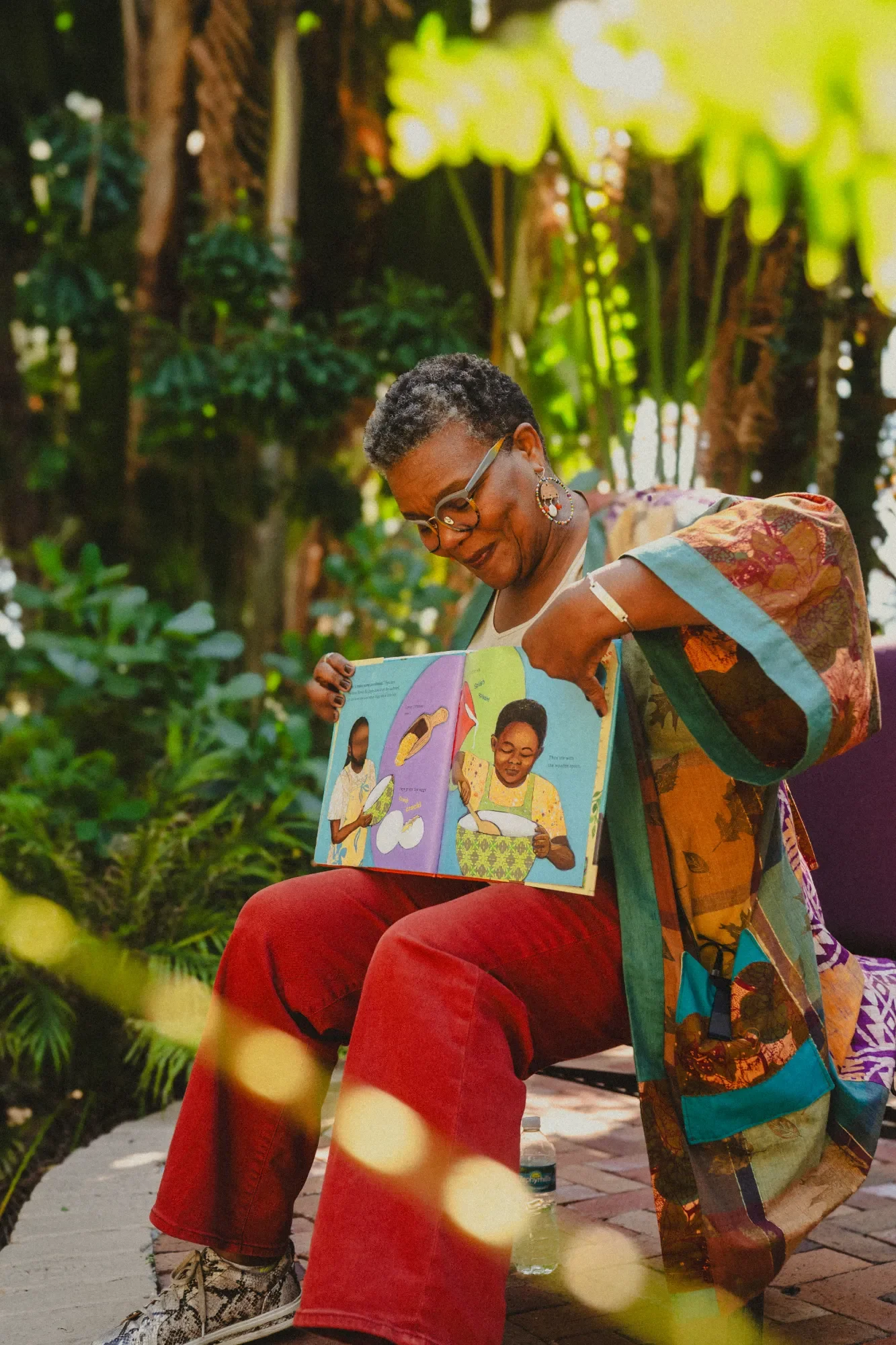 A person sitting outdoors in a lush, green setting reading a colorful children's book, wearing glasses, earrings, a patchwork jacket, and red pants.