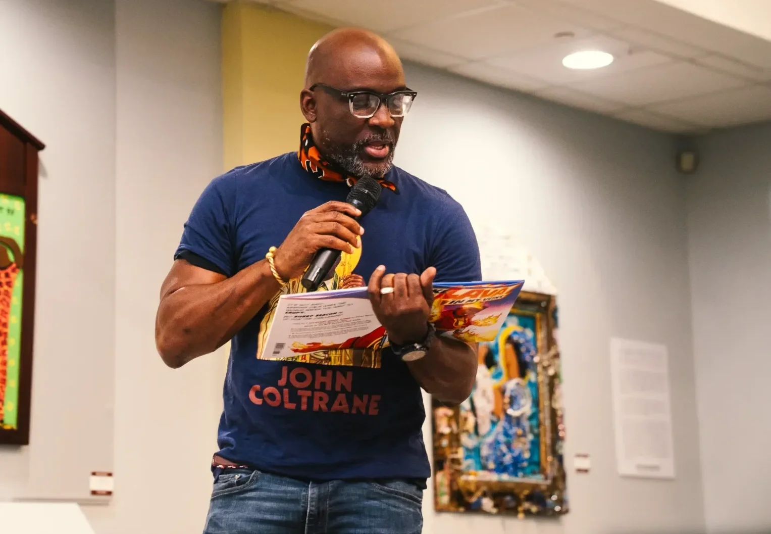 A man with glasses, wearing a dark blue t-shirt with 'John Coltrane' written on it, is holding a microphone and reading from a colorful pamphlet or magazine, standing indoors near a colorful artwork.