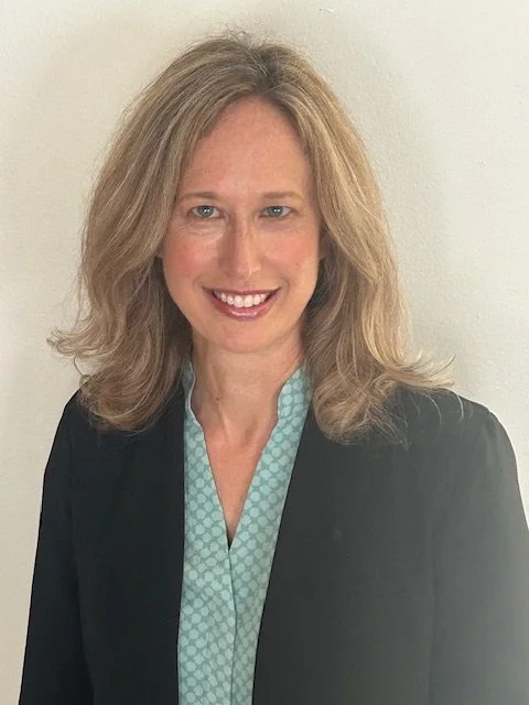 A woman with blonde hair wearing a black blazer over a light blue patterned blouse.