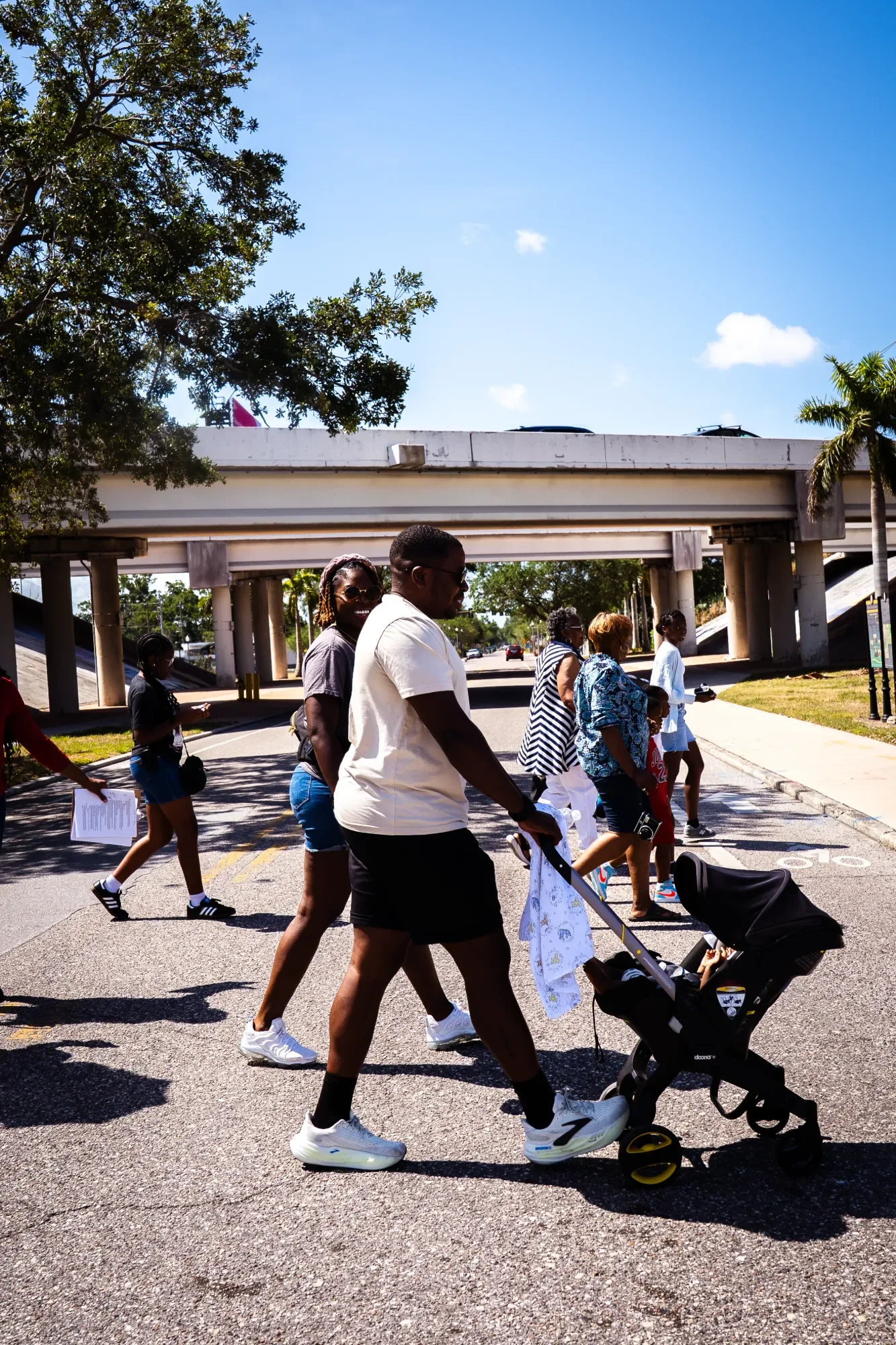 A diverse group of people crossing the street under a bridge on a sunny day, some pushing strollers, walking together, with trees and a blue sky in the background.