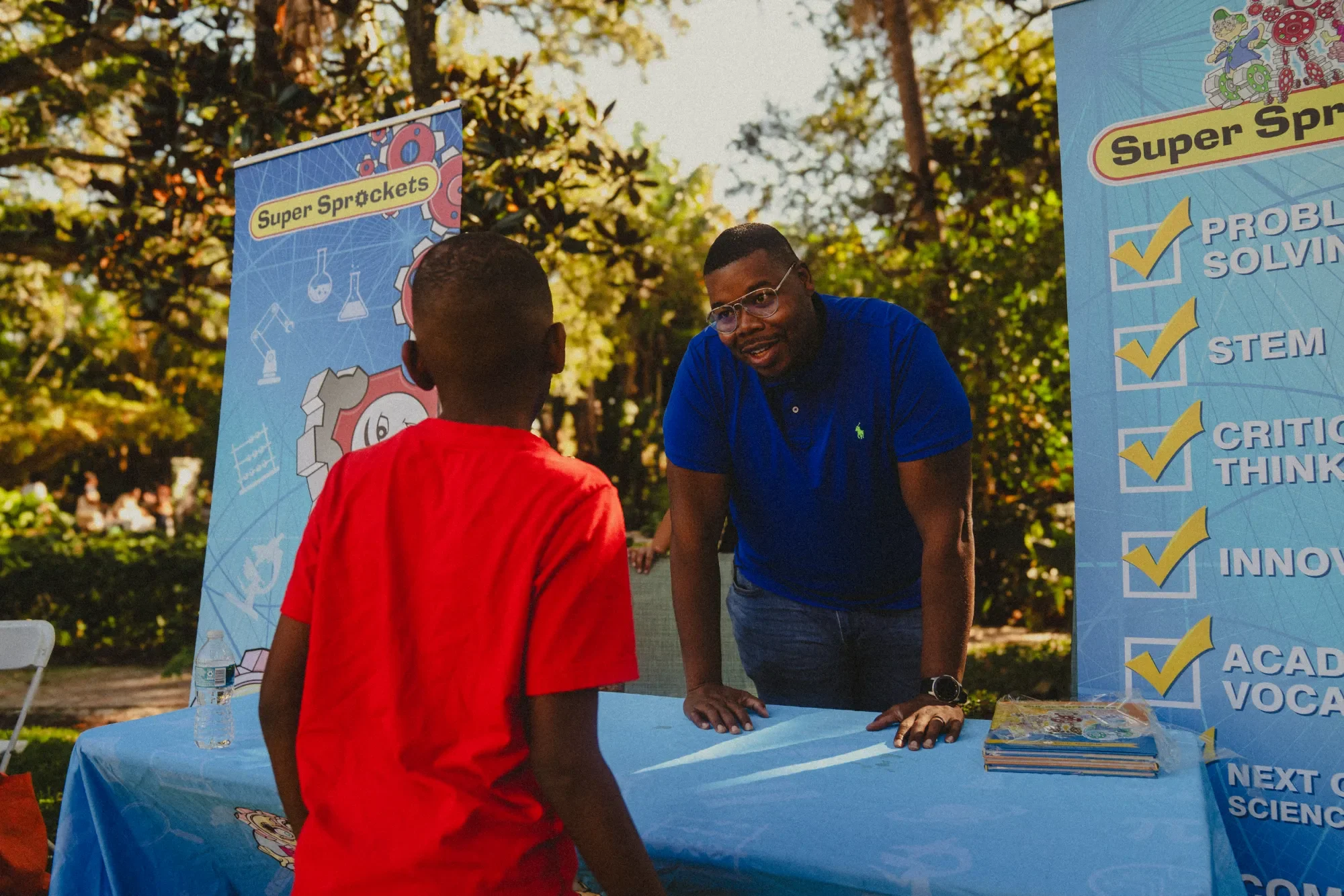 An outdoor science educational event with a man in a blue shirt talking to a young boy in a red shirt. There are informational banners with checkmarks and educational topics like STEM and critical thinking. The setting is a park with trees and sunlight.