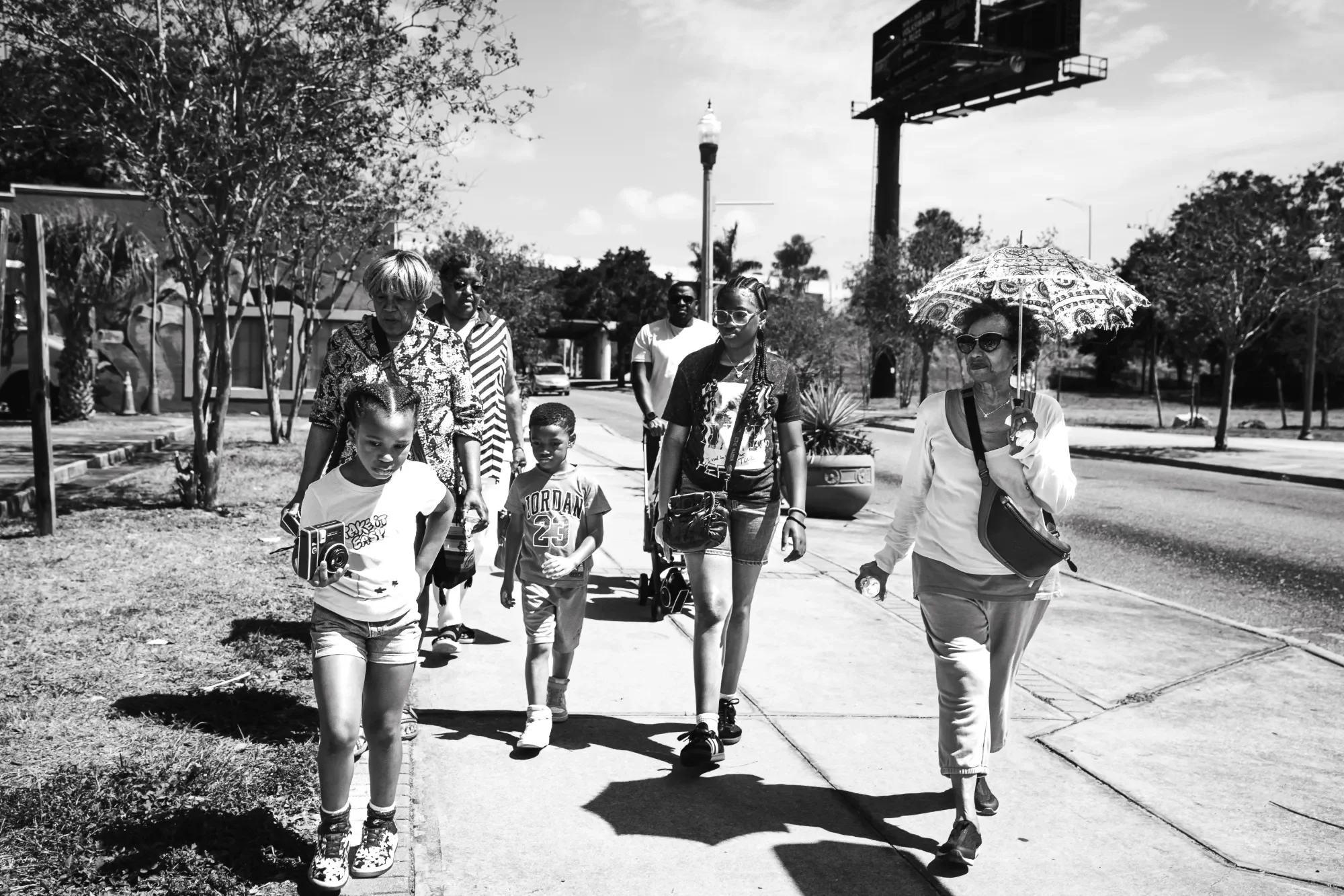 A diverse group of people, including two young children and several adults, walking on a sidewalk in a sunny outdoor setting with trees and a large billboard in the background. One woman is holding an umbrella to shield herself from the sun.