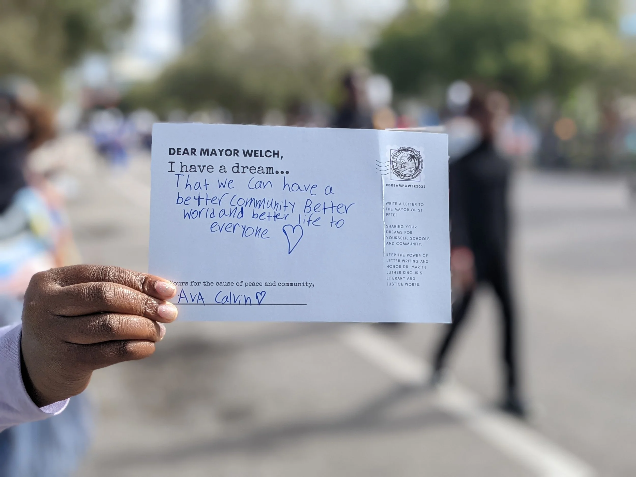 Hand holding a handwritten letter addressed to Mayor Welch with a heart drawing, outdoors on a sunny day with blurred people in the background.