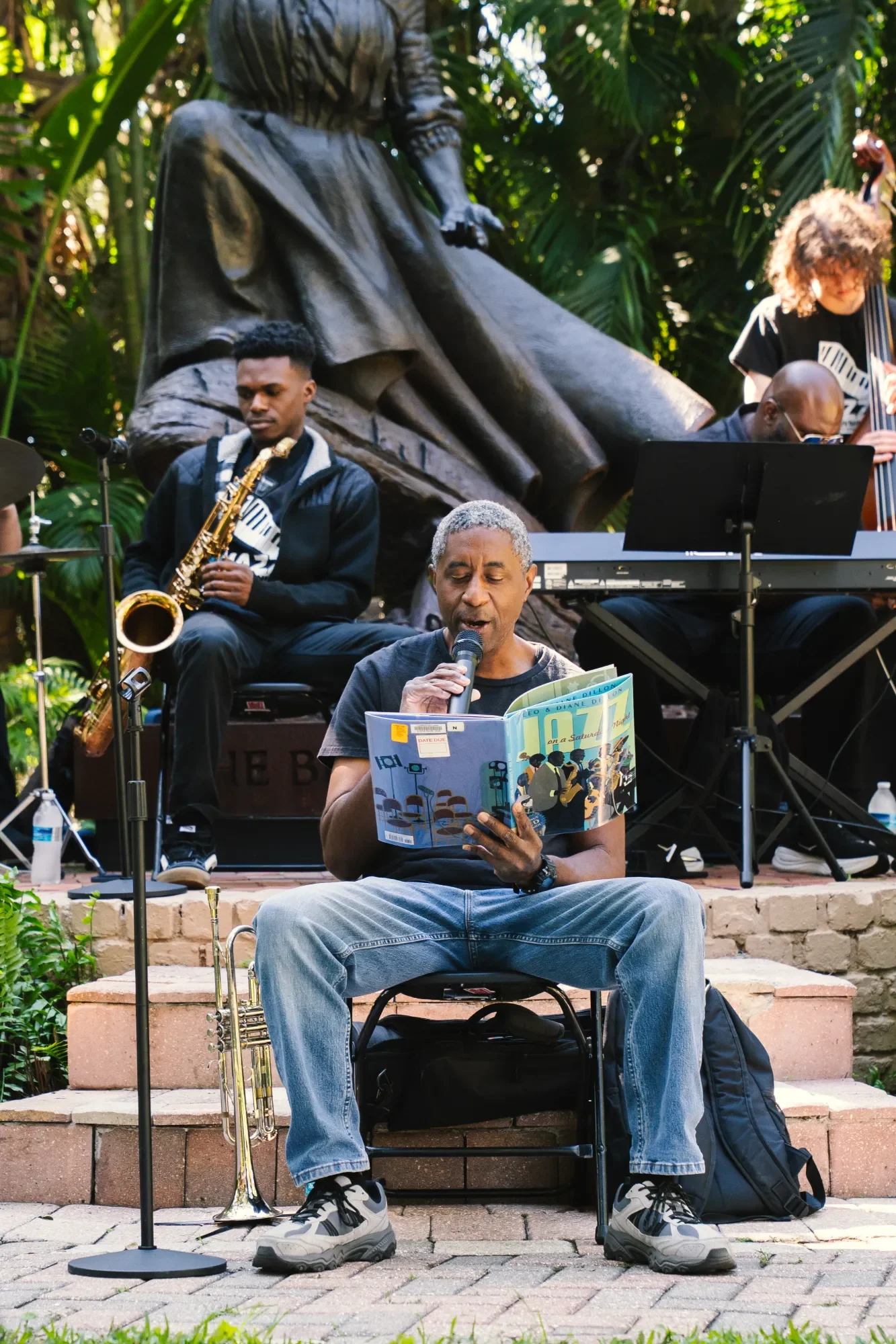 A man sitting on a chair reading a book and singing into a microphone outdoors, with a saxophone on the ground beside him. Behind him, a music band with musicians playing instruments is set up on stone steps. A large bronze statue of a woman is in the background among lush green plants.