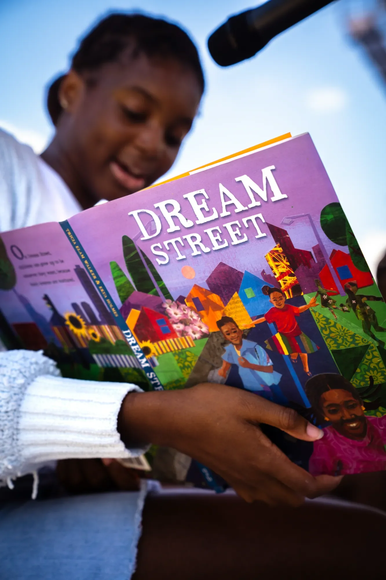 A person is holding and reading a colorful booklet titled 'Dream Street' with illustrations of children and buildings on the cover, outside under a clear blue sky.