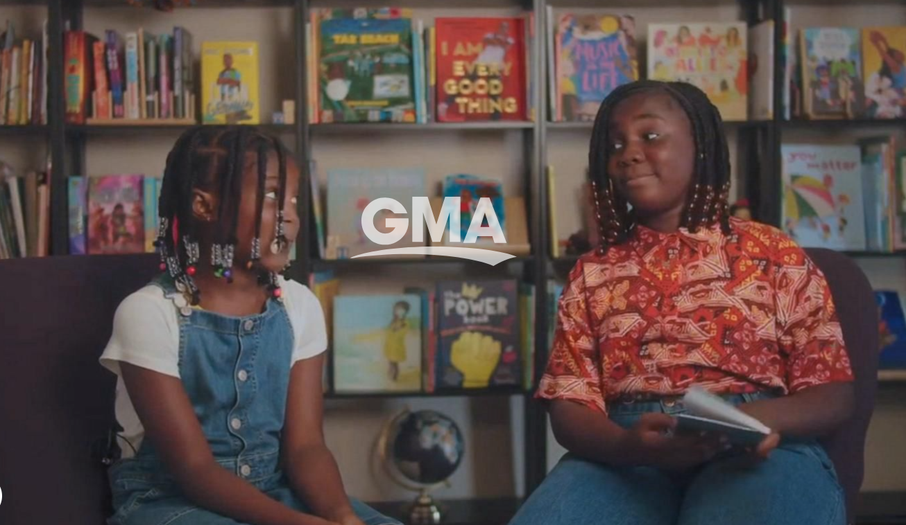Two young girls with braided hair and bead accessories sitting in a library, engaged in conversation. Bookshelves filled with colorful children's books are in the background.