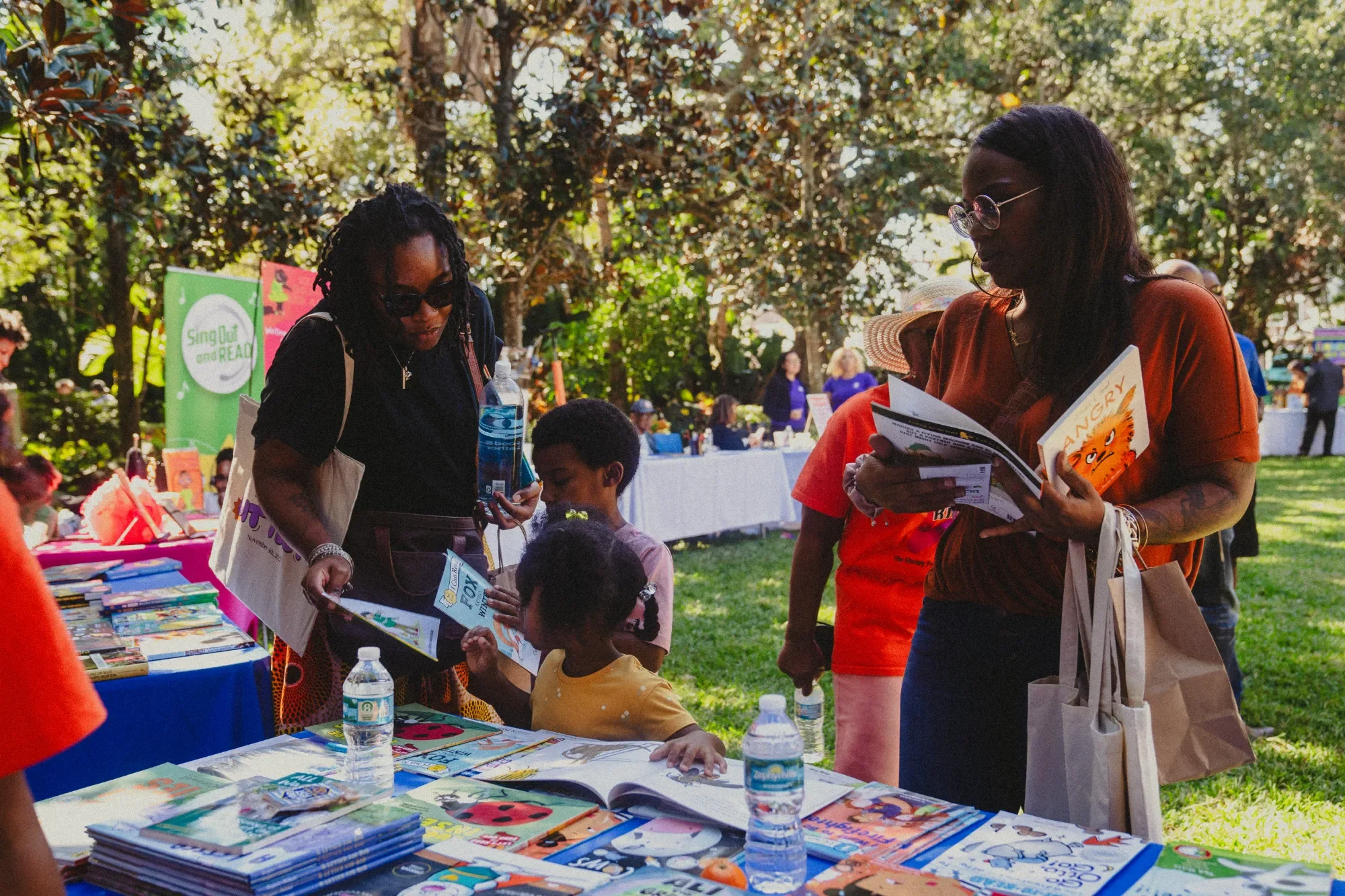 People at a table filled with books, magazines, and water bottles outdoors at a daytime event with people and trees in the background.