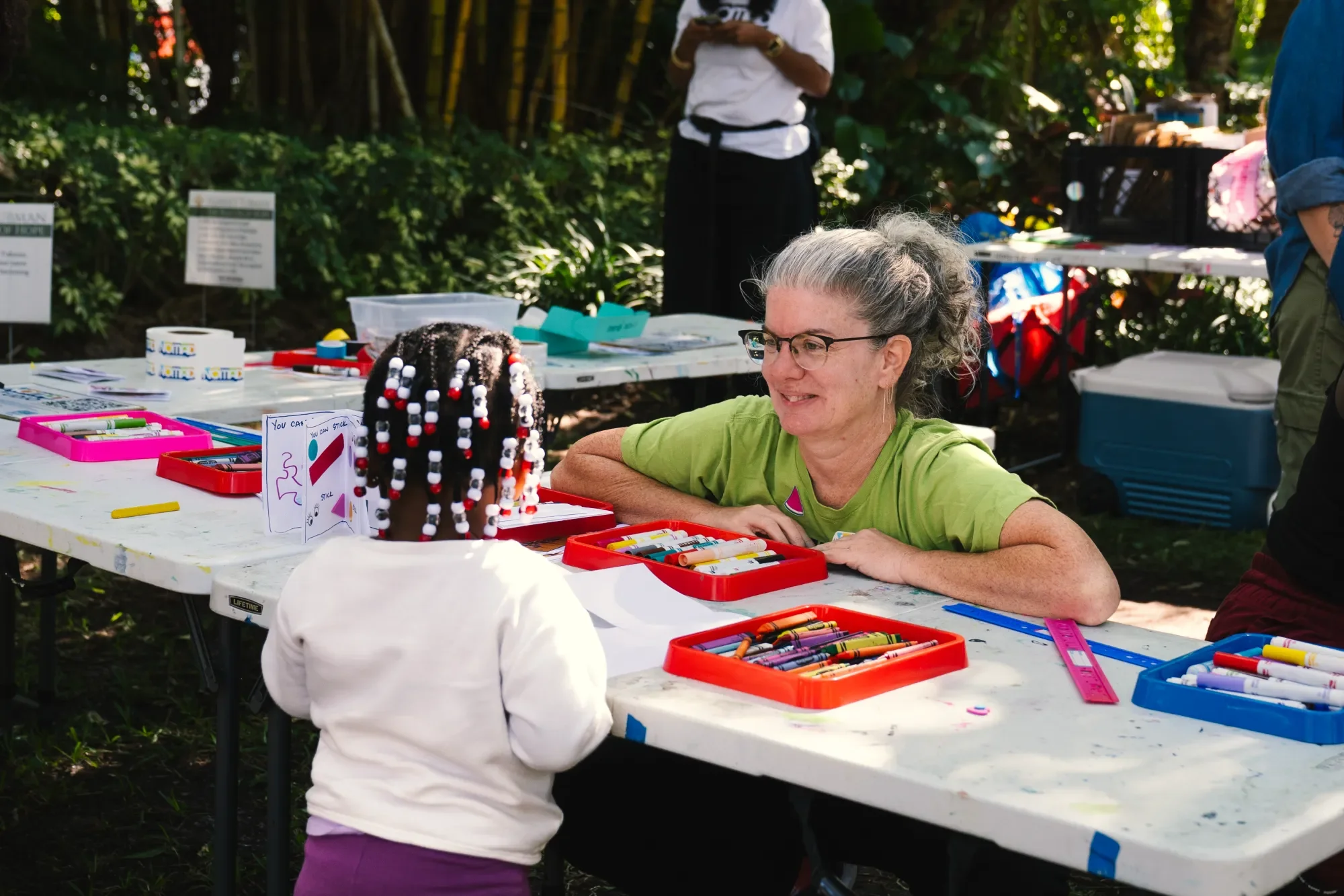 A woman with glasses and gray hair, wearing a green shirt, smiling at a young girl in a white shirt and purple pants, who is holding a craft project with beads and letters. They are at a table outdoors with art supplies, in a green, wooded area.