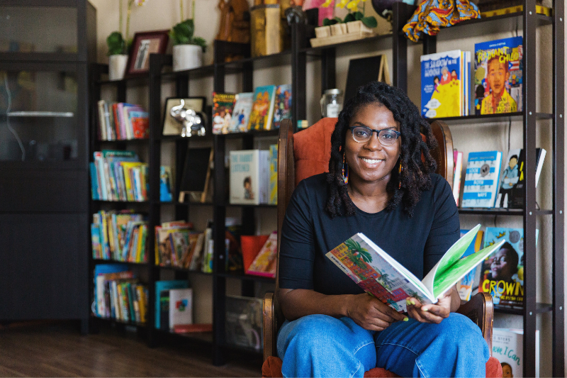 A woman sitting on a chair in front of bookshelves filled with colorful children's books, smiling and holding an open book.