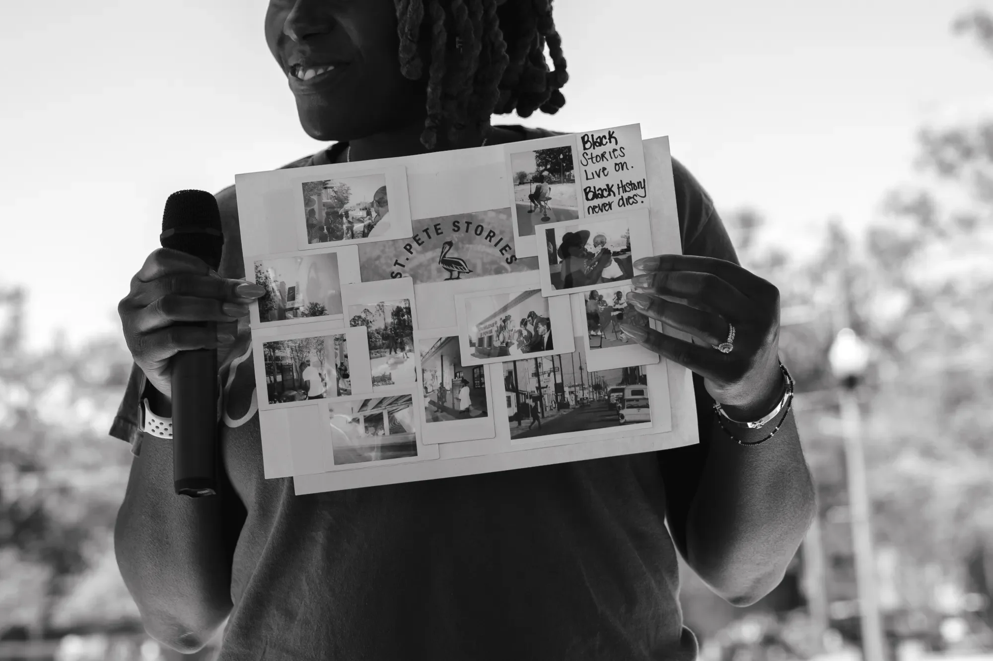 A woman with dreadlocks holding a bulletin board with photos and notes, smiling and standing outdoors, with a microphone in her hand.