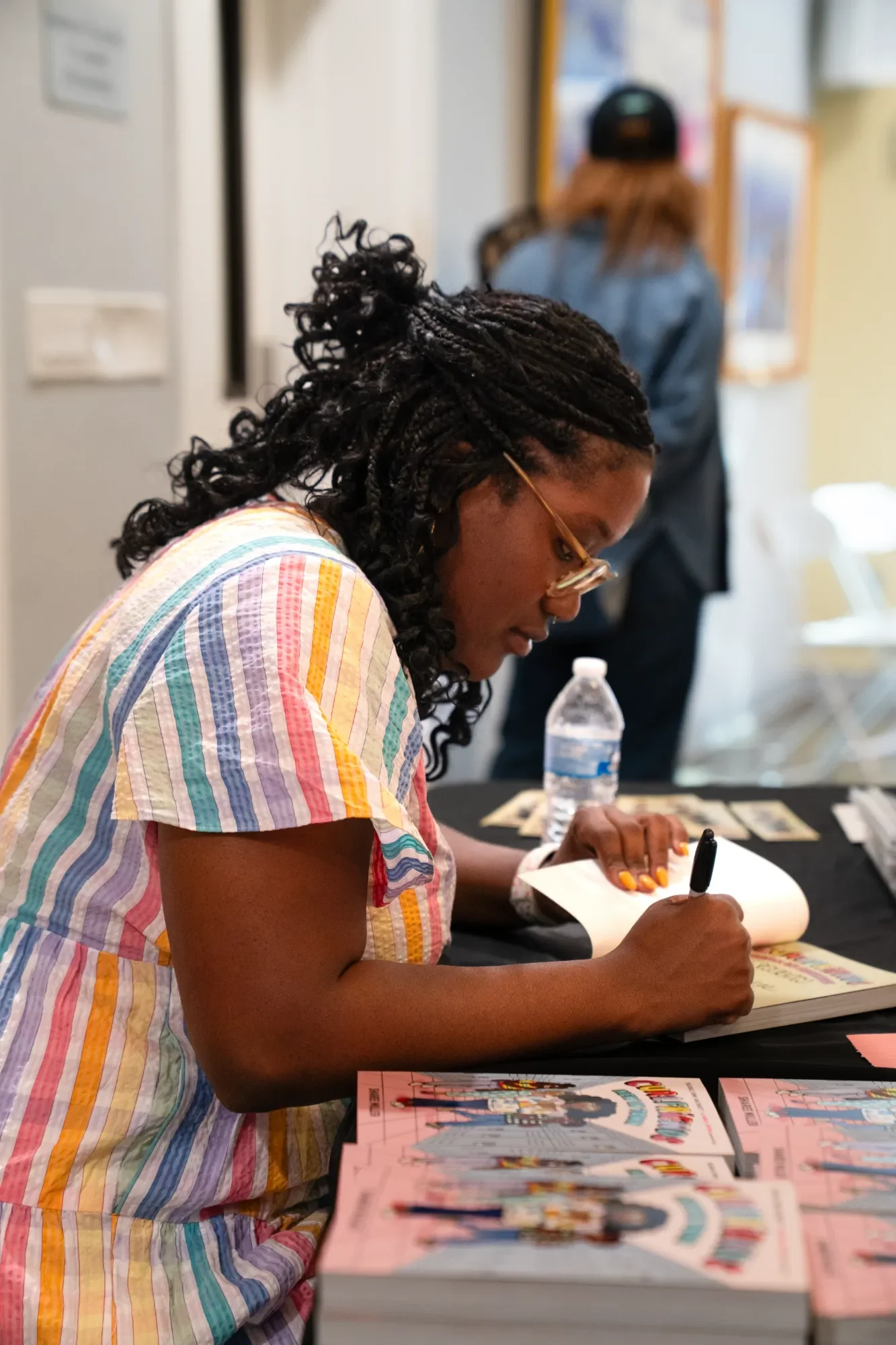 A woman with styled black dreadlocks and glasses, wearing a colorful striped shirt, is sitting at a table, signing a book with a black marker. On the table are more books and a water bottle. In the background, another person with long hair, a black cap, and a denim jacket is standing and facing away. The setting appears to be a book signing event or a meet-and-greet.