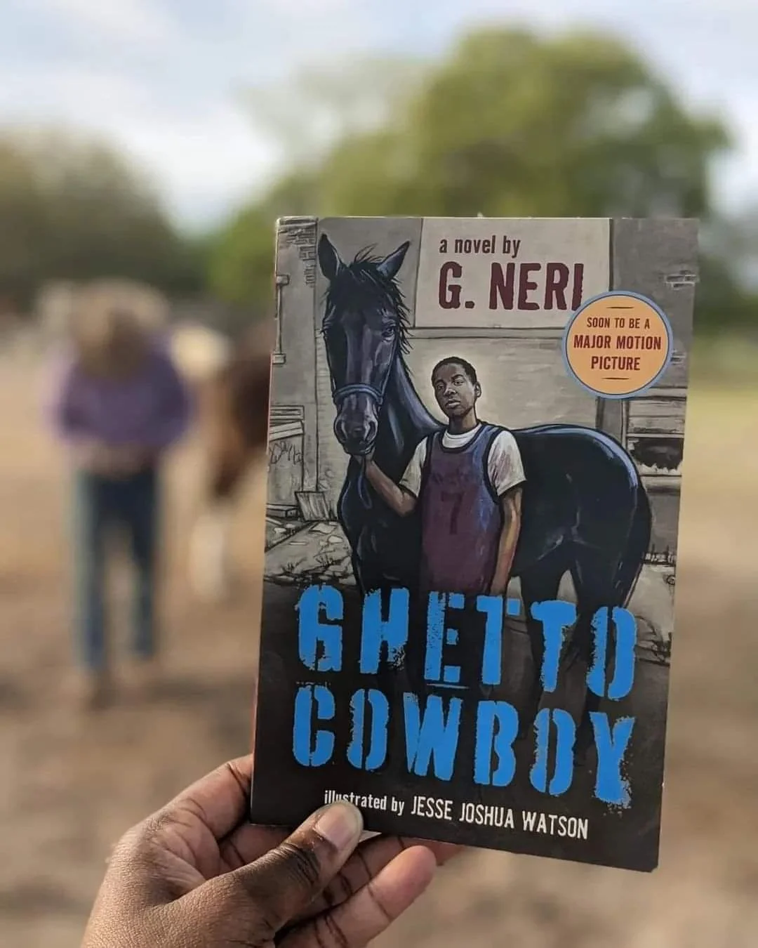A person holds a book titled 'Ghetto Cowboy' with an illustration of a young man and a black horse on the cover. The background shows two blurred figures and greenery.