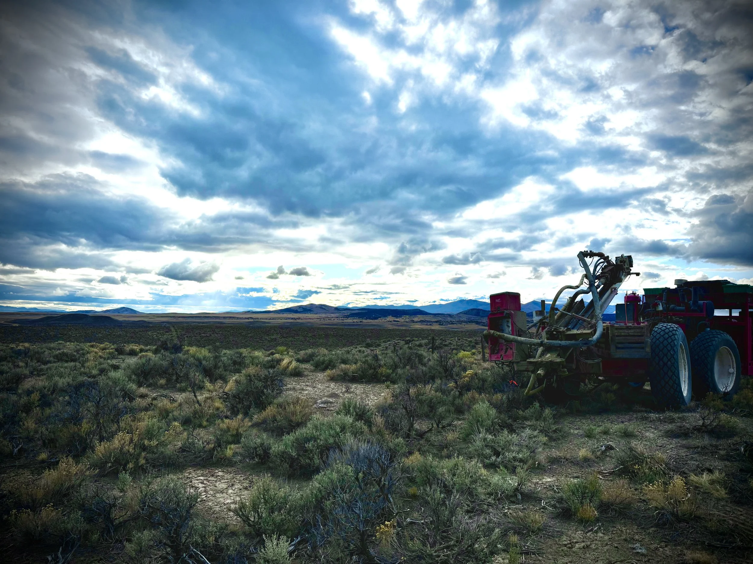 A large open landscape with scrubby bushes and distant mountains under a partly cloudy sky, with heavy machinery on the right side of the scene.