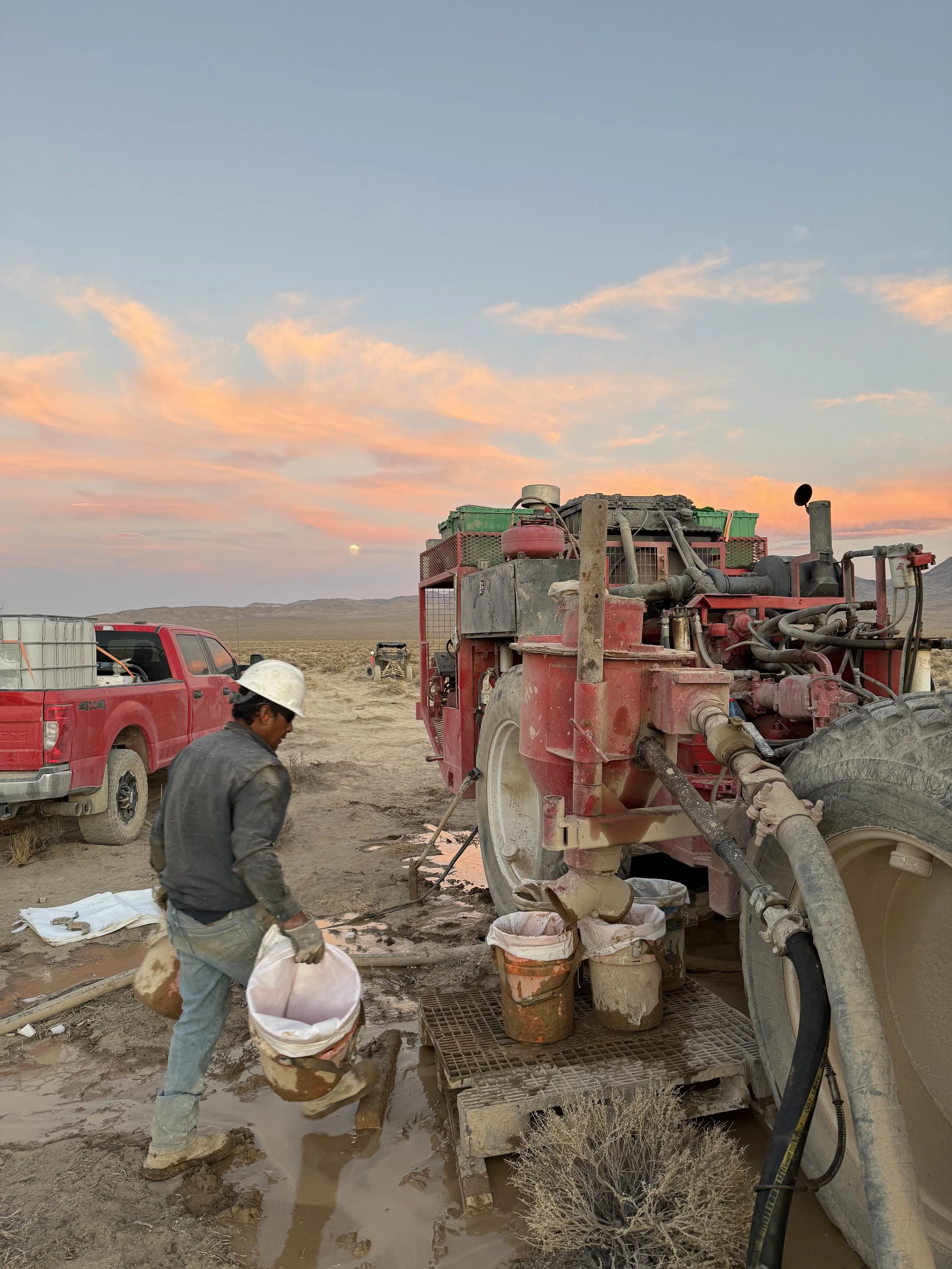 A worker in a hard hat and gloves operating heavy machinery in a desert at sunset, with trucks and equipment in the background.