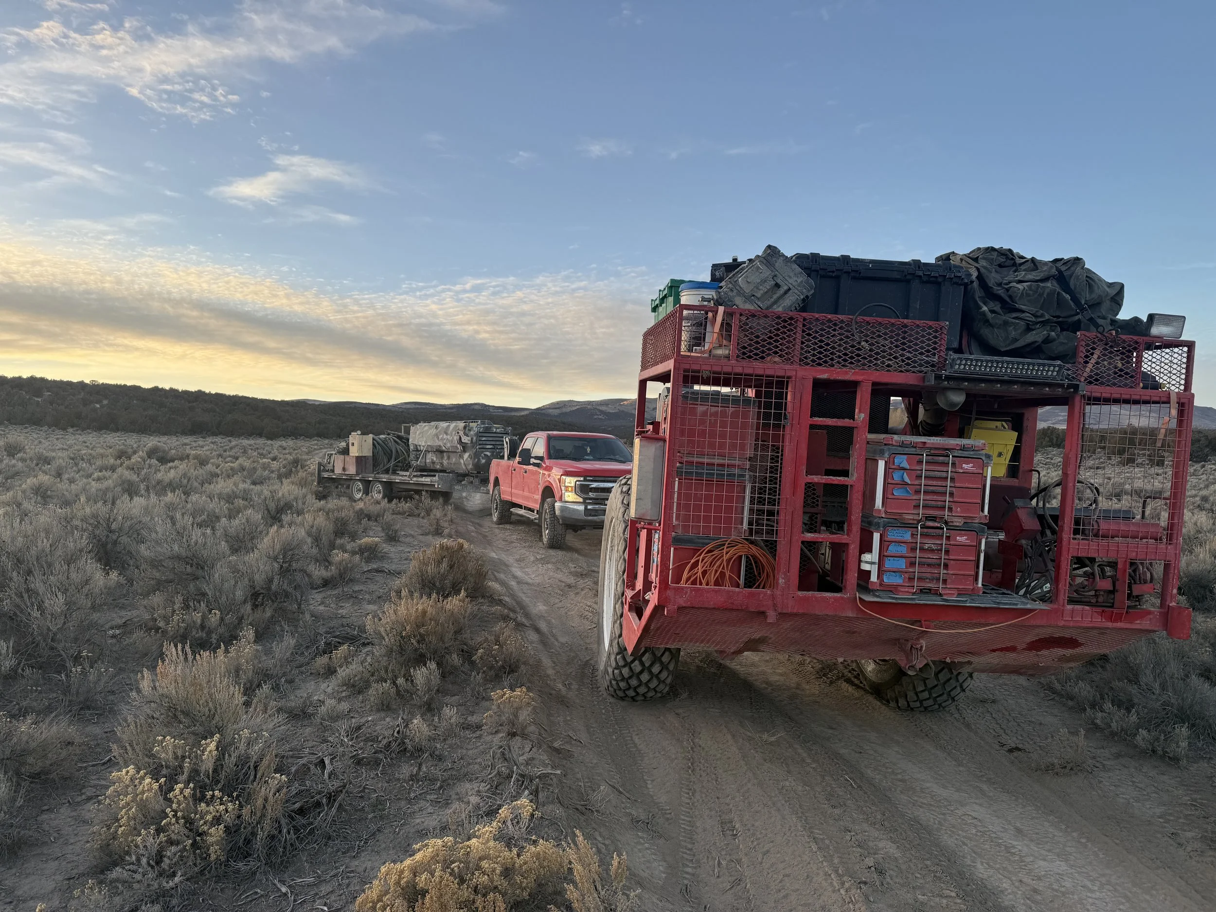 Red utility trailer filled with equipment and supplies, being pulled by a red pickup truck, on a dirt trail in a desert landscape with sparse bushes and a colorful sky at sunset.