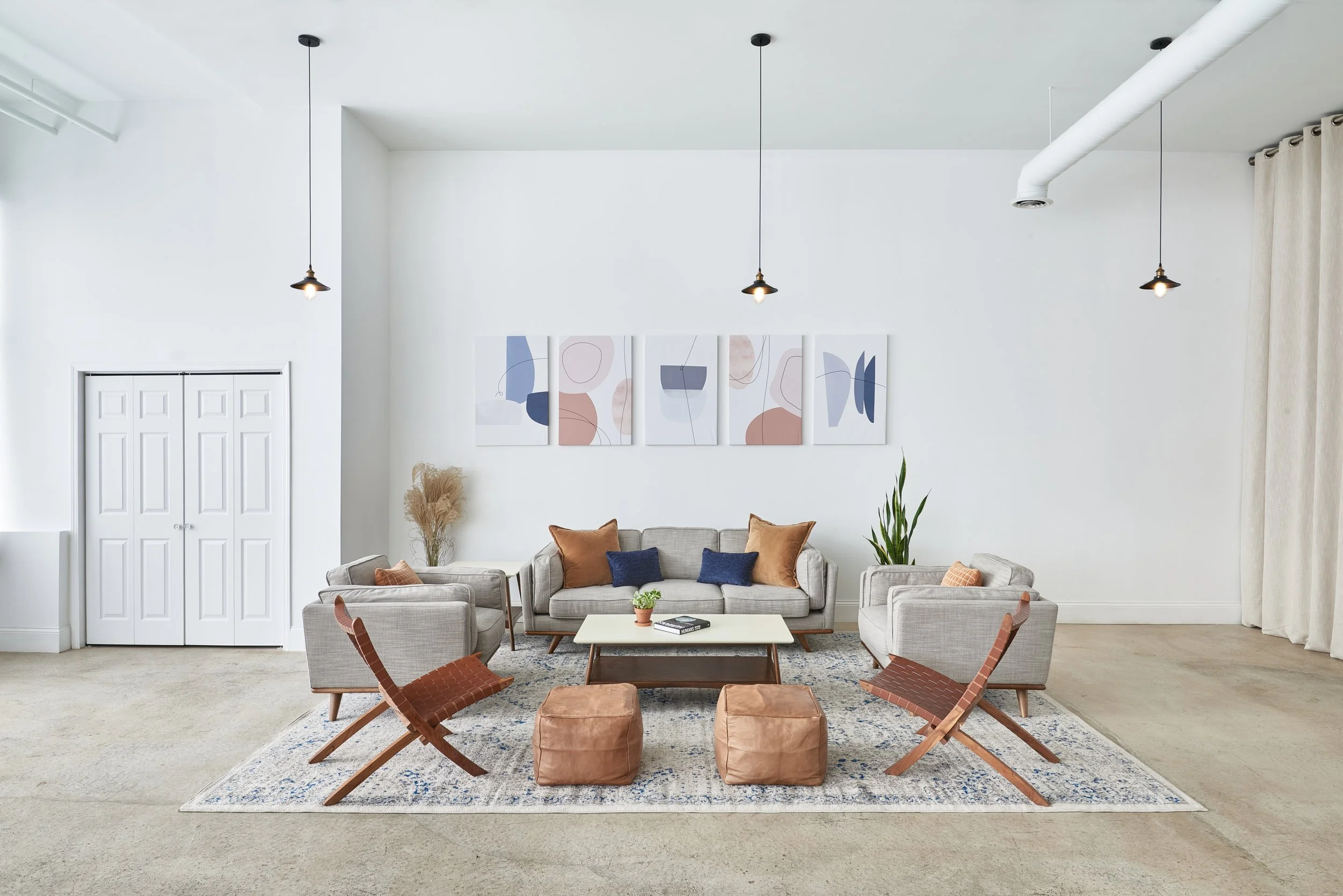 Modern living room with beige sofas, brown accent pillows, matching chairs, a coffee table, potted plants, abstract wall art, and hanging ceiling lights.