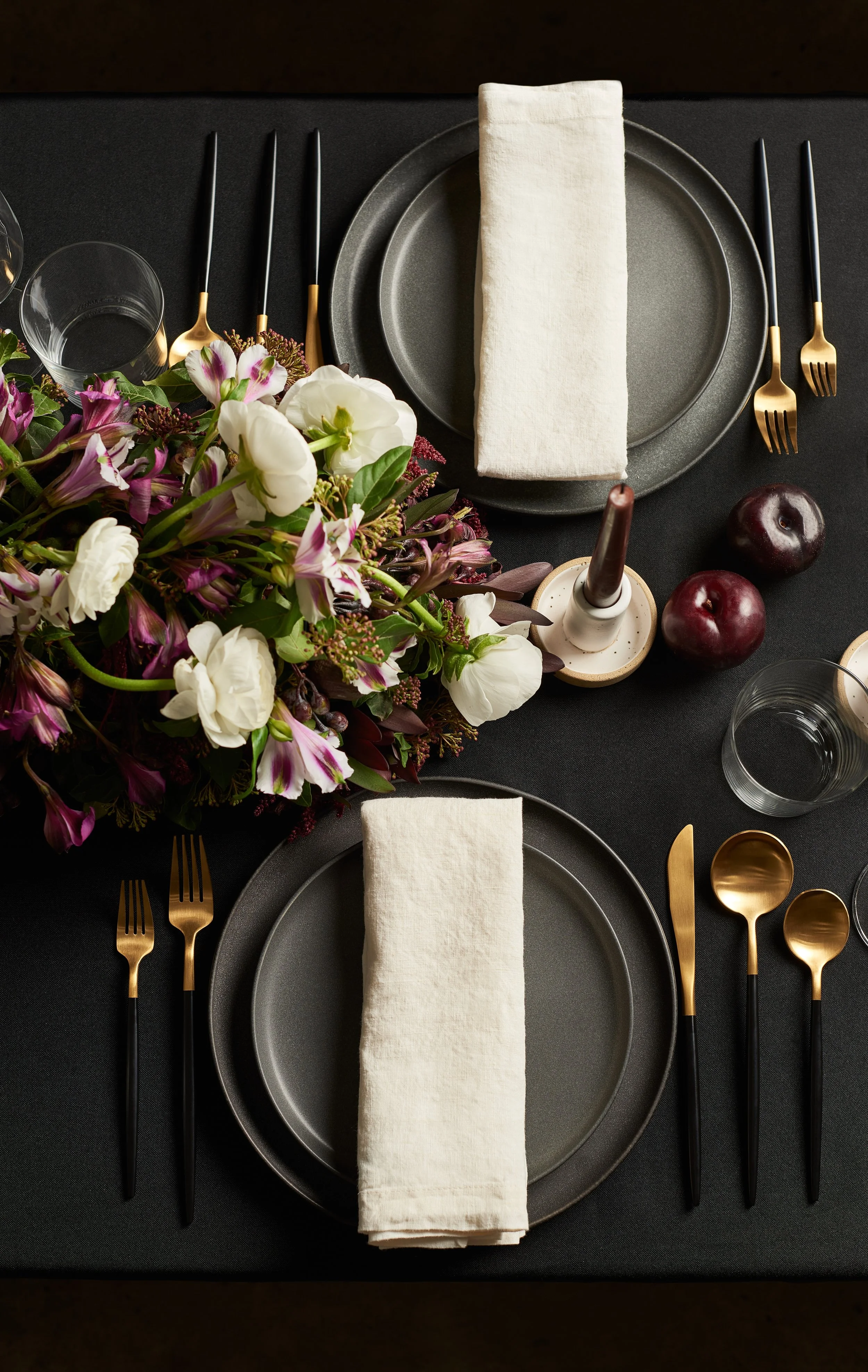 Elegant dining table setting with black plates, gold and black utensils, white cloth napkins, a floral centerpiece, two apples, and glasses on a black tablecloth.