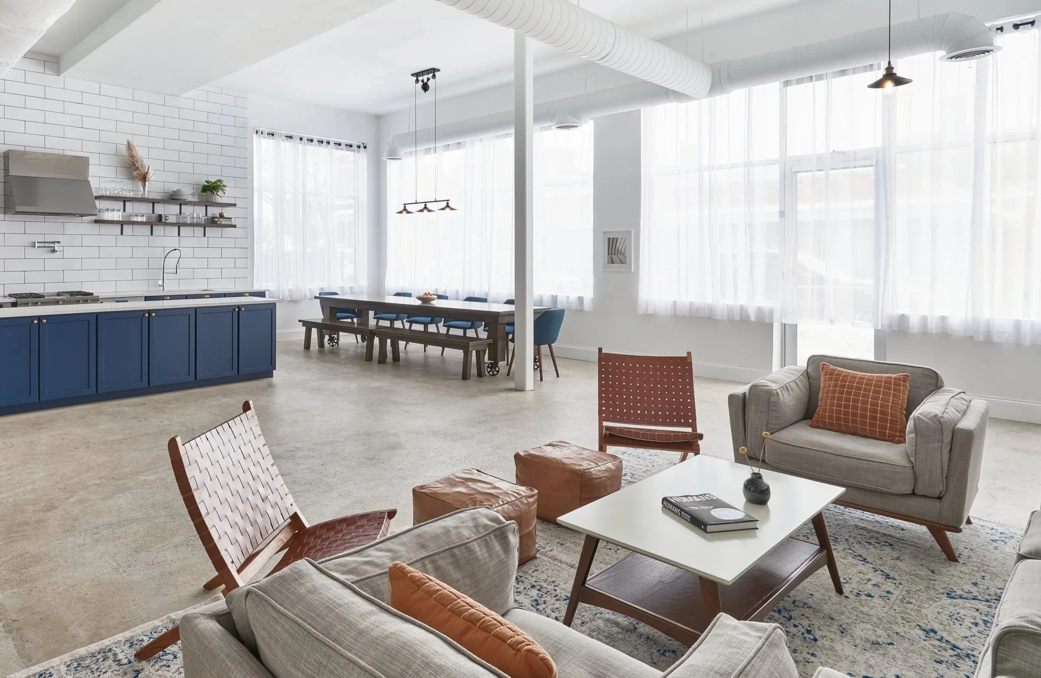 Bright open-concept living room and kitchen with large windows, white walls, and eclectic furniture including beige sofas, a white coffee table, leather ottomans, and a blue kitchen island.