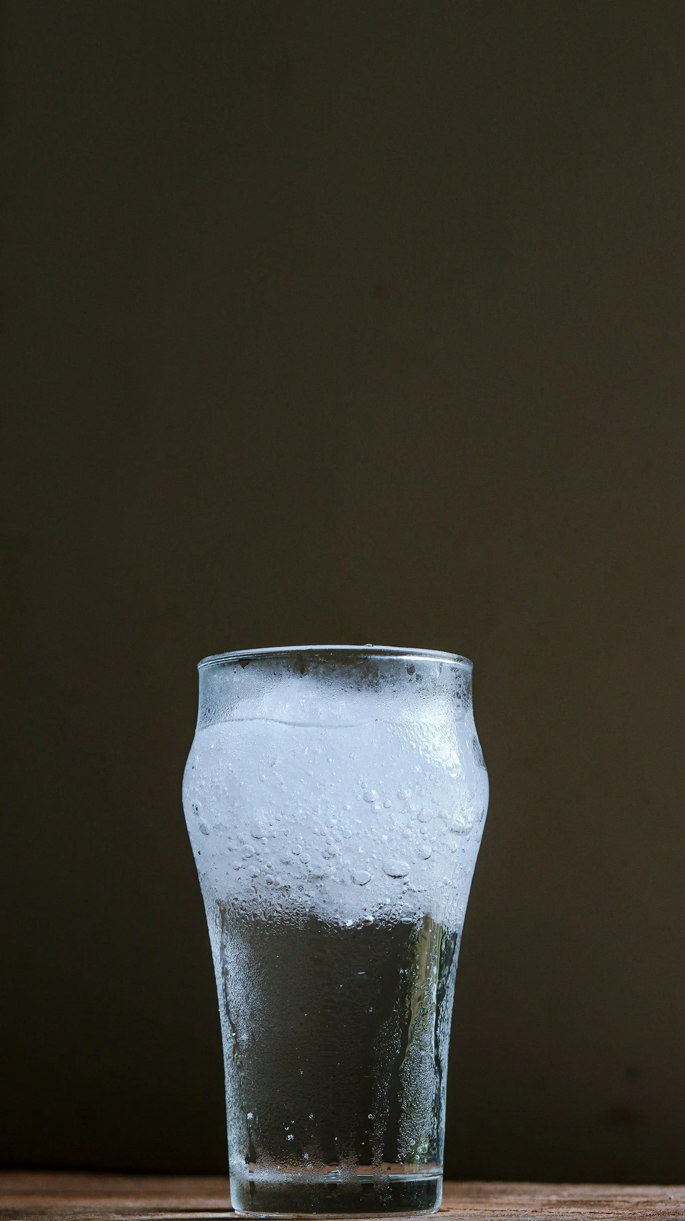 A tall glass of ice water with condensation on the outside, sitting on a wooden surface against a dark background.