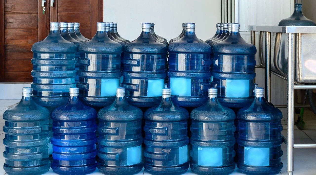 Stacked large blue water bottles with caps, arranged in three rows, indoors near a kitchen or utility area.