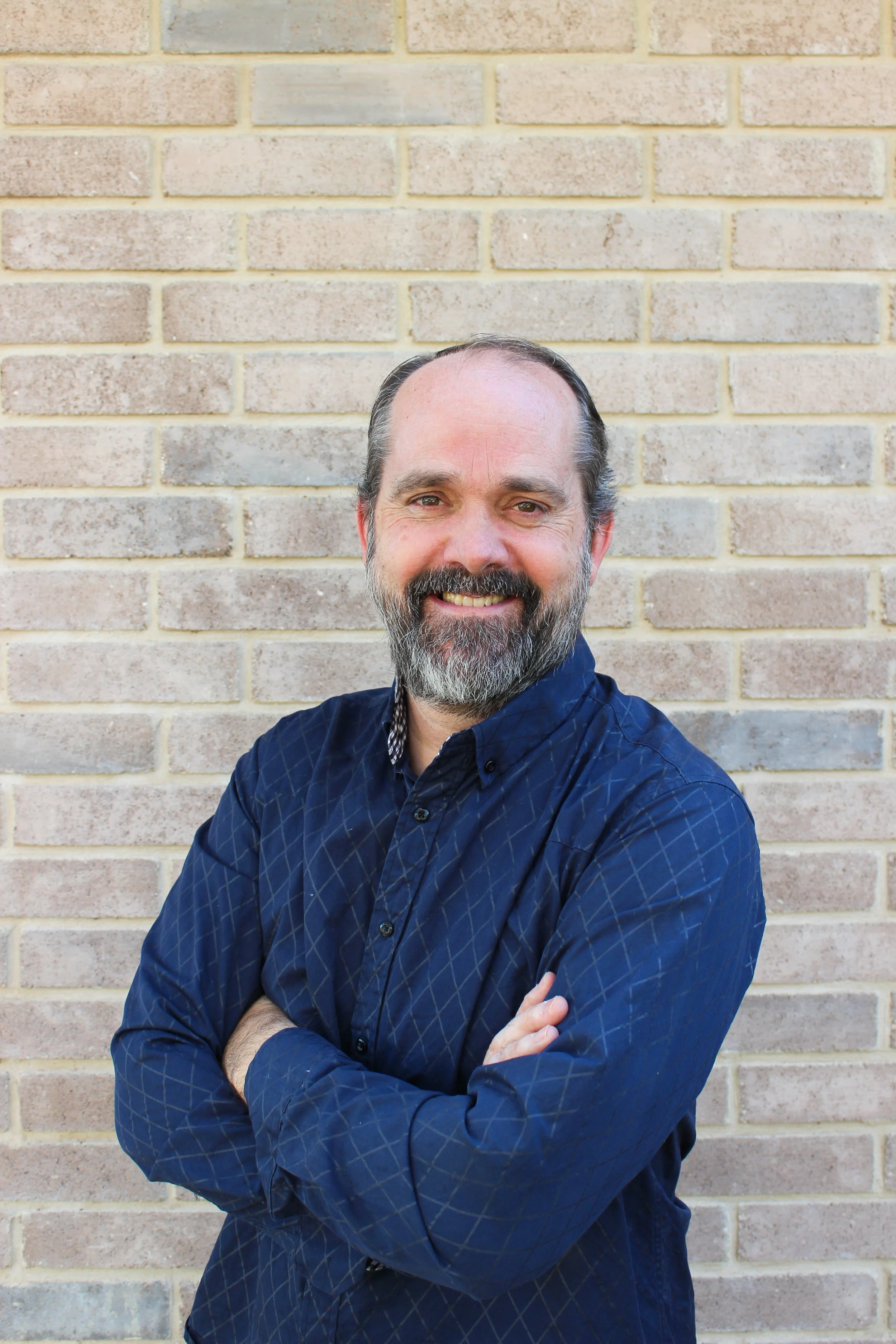 A smiling middle-aged man with a beard wearing a dark blue button-up shirt with a checkered pattern, standing against a beige brick wall with his arms crossed.