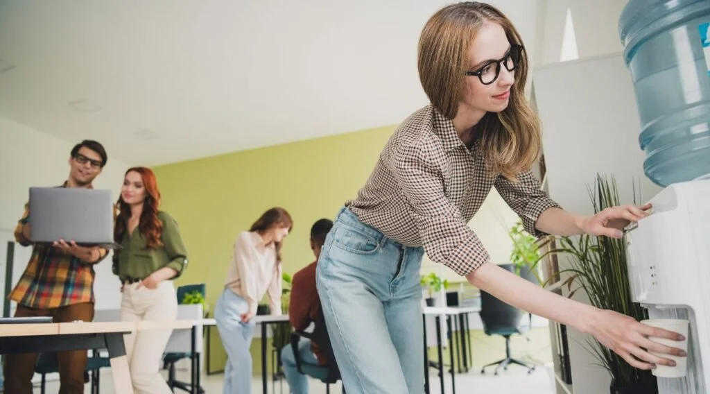 People in an office, with a woman filling a water bottle from a water cooler in the foreground, and three other individuals working or talking in the background.