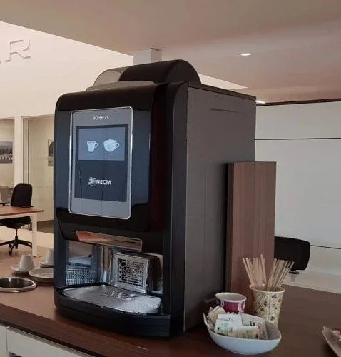 A coffee vending machine in a modern office break room with cups, stir sticks, and snacks nearby.