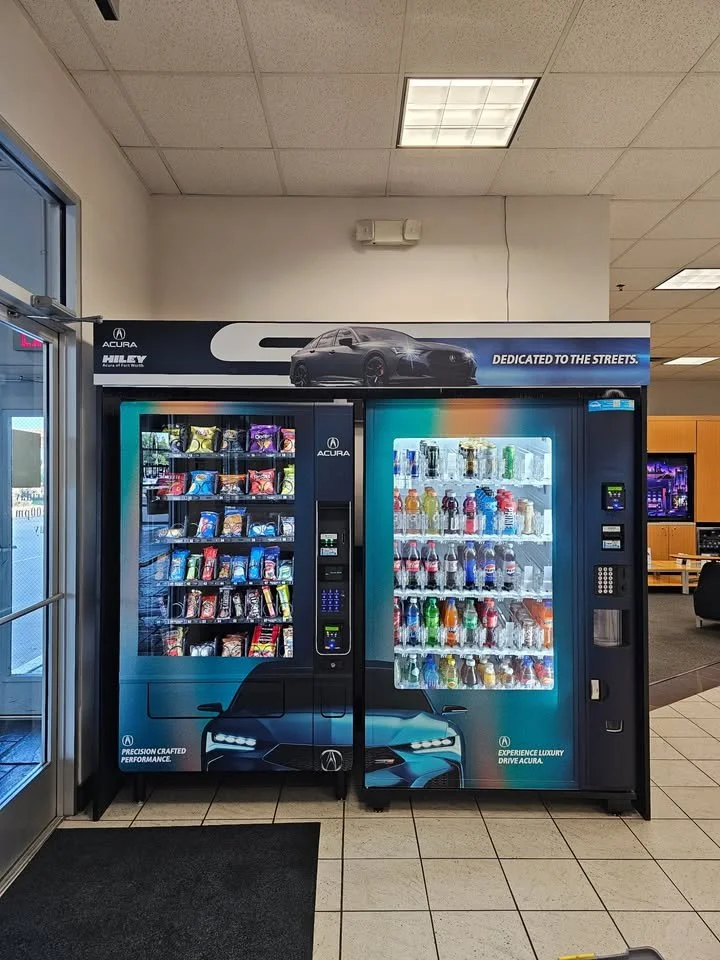 Two vending machines side by side inside a building, one stocked with snacks and the other with beverages, next to an entrance door.