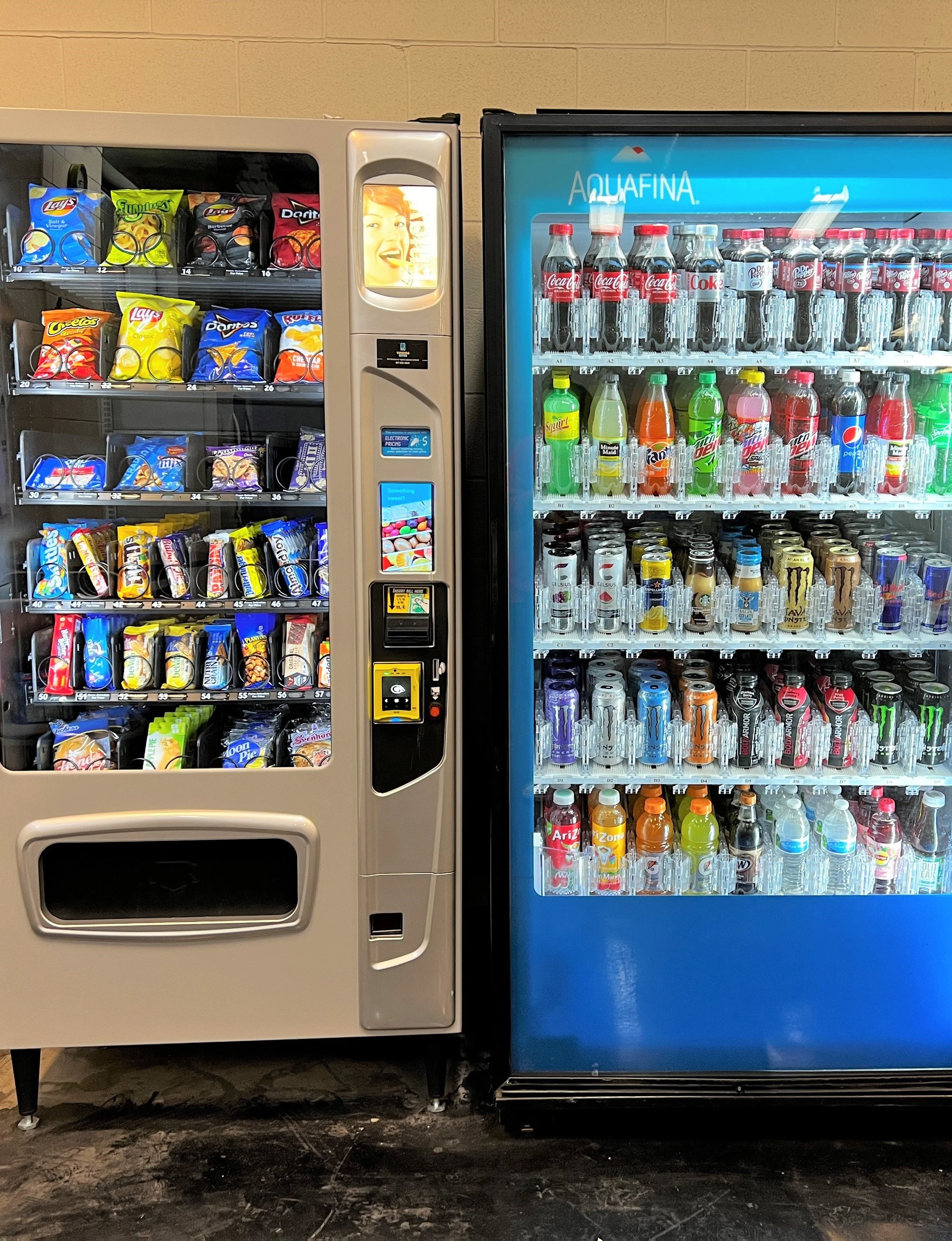 Vending machine filled with snack foods and a soda vending machine stocked with various beverages, including Coca-Cola, Sprite, Mountain Dew, and Monster energy drinks, against a beige wall.