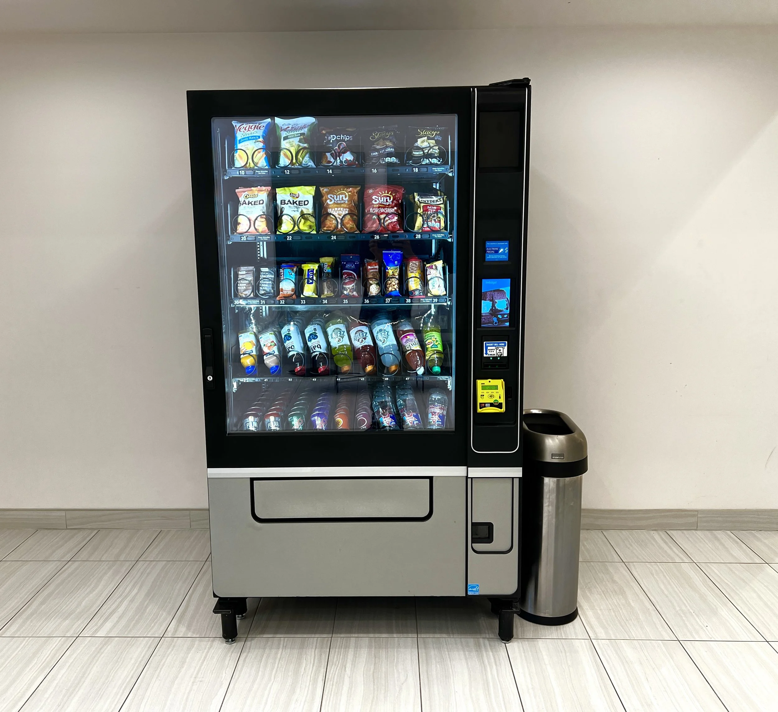 A vending machine stocked with various snack chips and beverages, situated against a beige wall with tiled flooring.