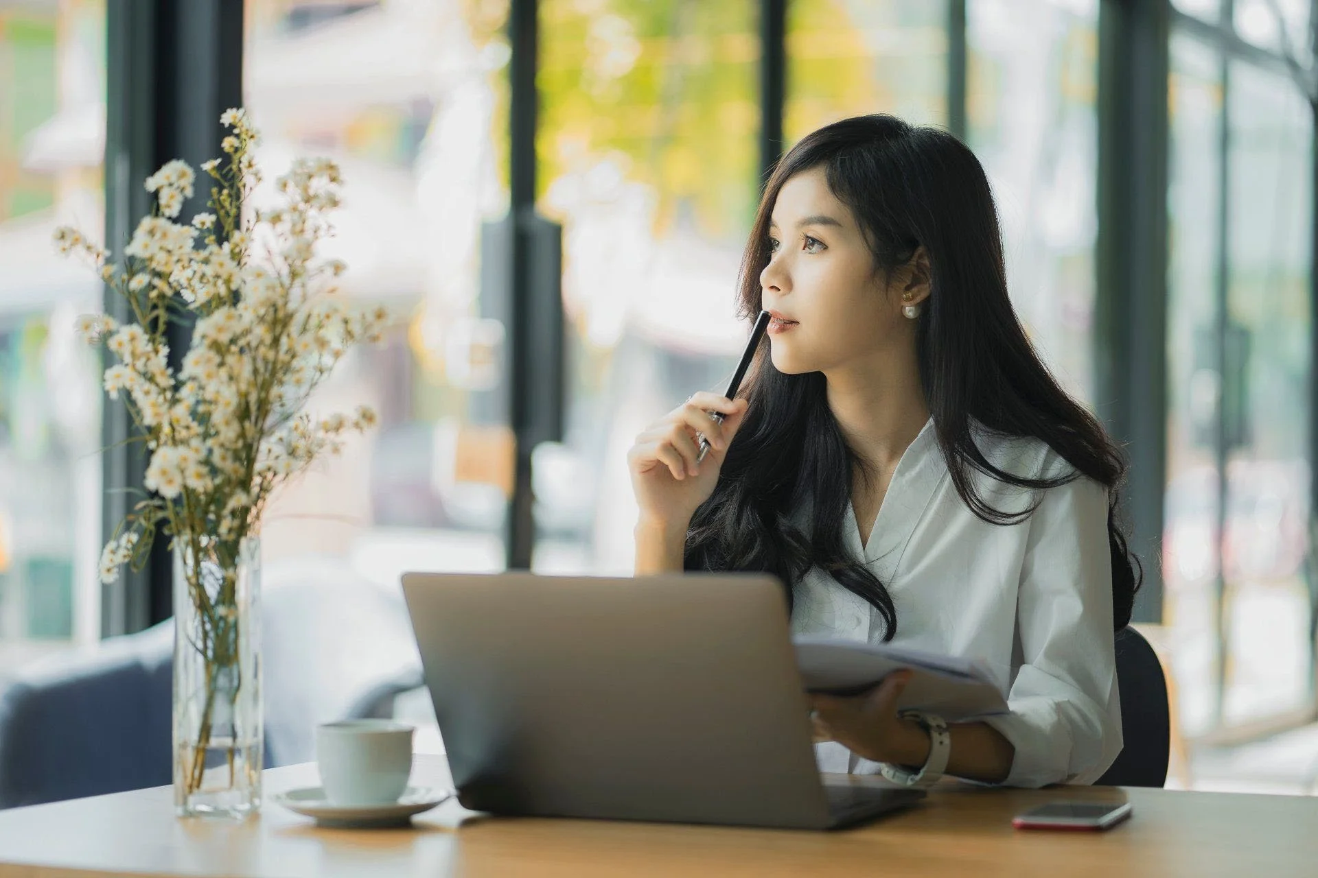 A woman with long dark hair wearing a white shirt is sitting at a table in a well-lit room with large windows. She is holding a pen to her lips and looking thoughtfully out the window. On the table, there is a laptop, a white coffee mug, a smartphone, and a tall vase with white and yellow flowers.