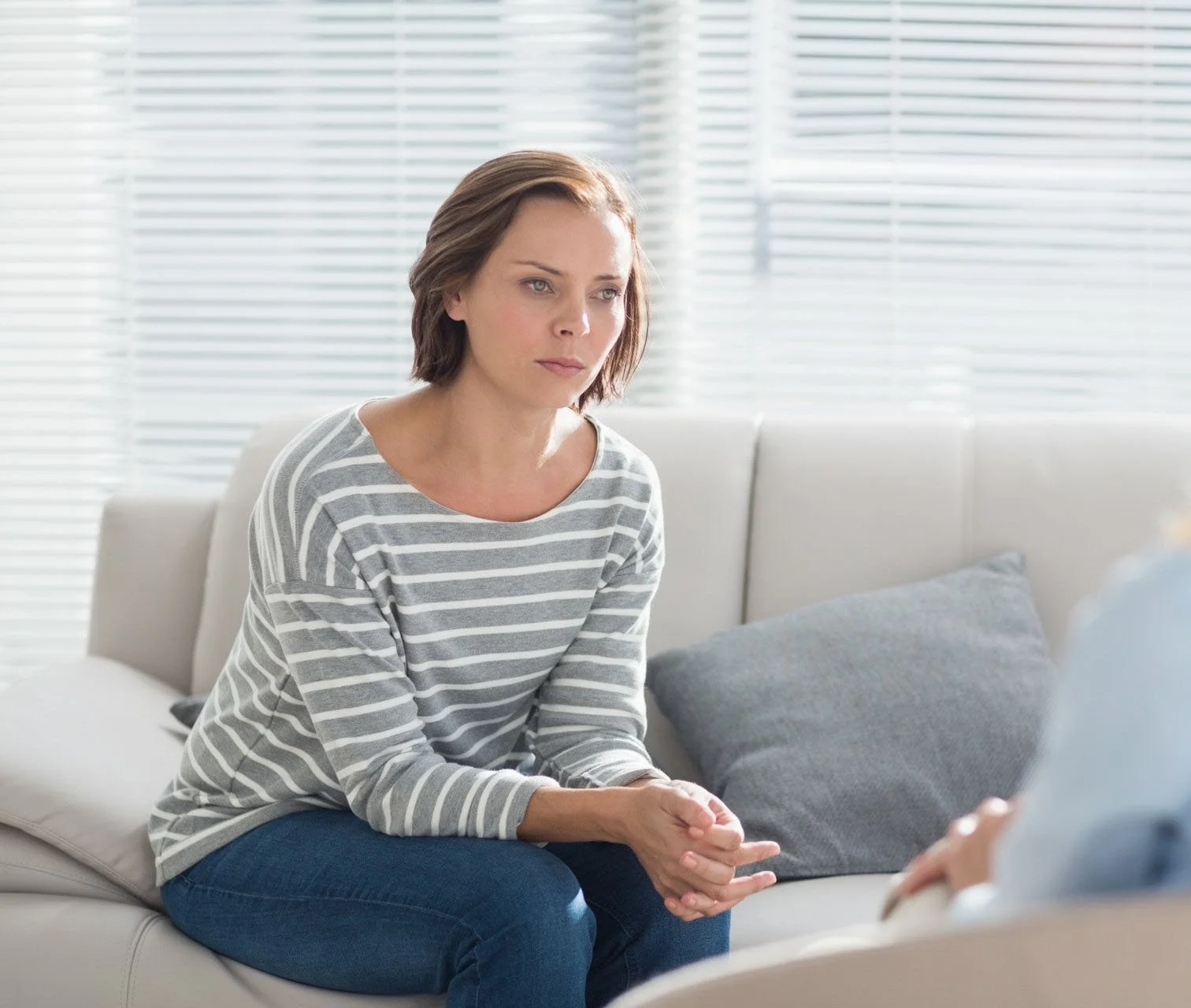 A woman with short brown hair wearing a striped gray and white shirt, sitting on a white couch with gray cushions, talking to someone outside the frame, in a room with white blinds and natural light.