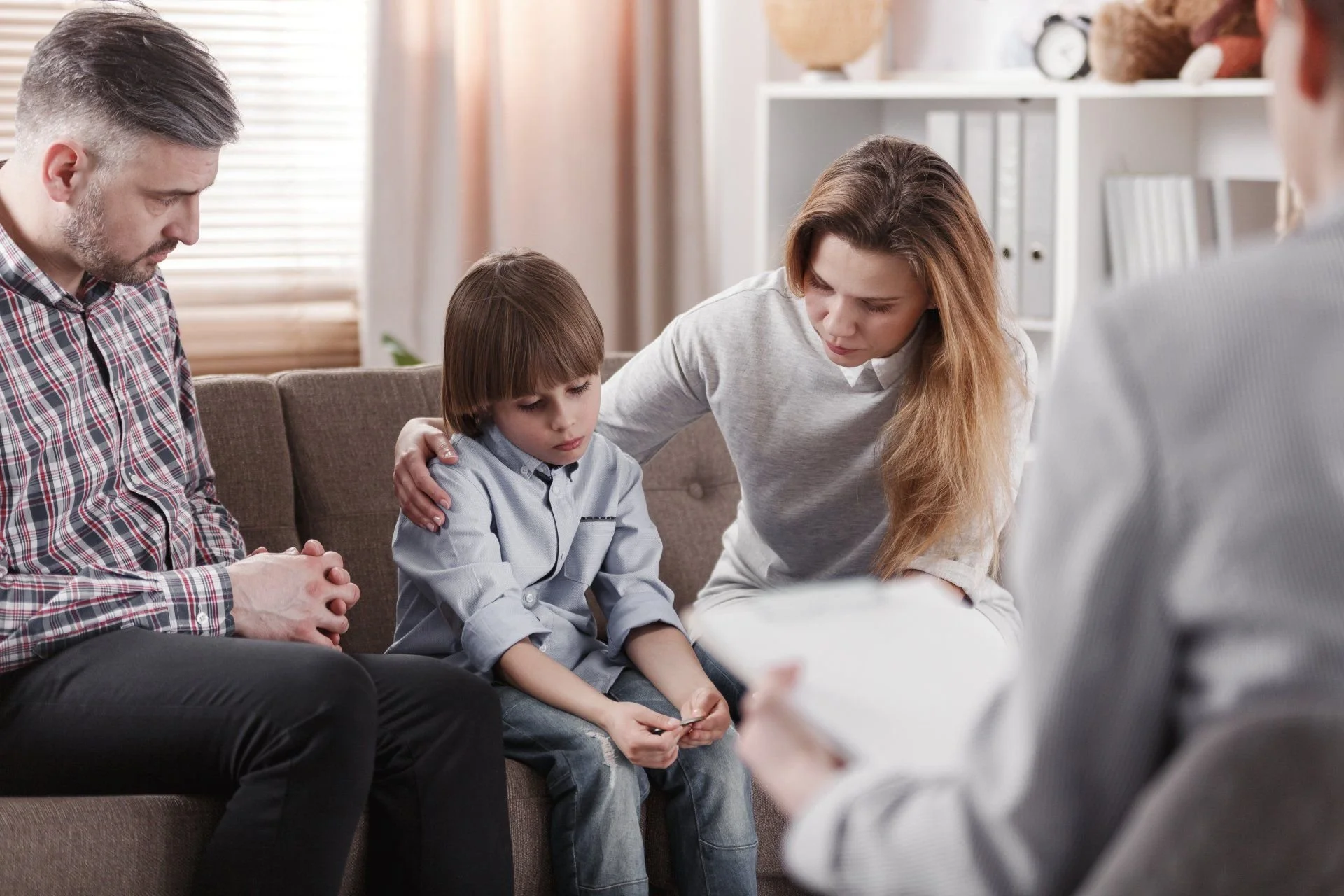 A family sitting on a sofa in a living room, with a woman and a young boy in the center, experiencing a moment of emotional support or counseling from an unseen professional.