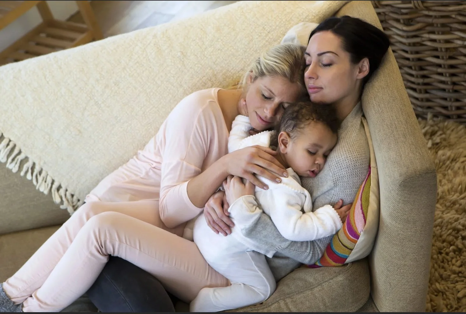 Two women and a young girl cuddling and sleeping together on a beige couch in a cozy home setting.