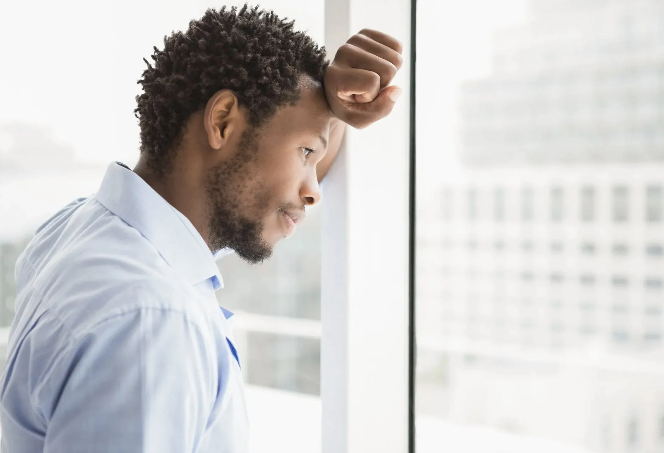 A man with curly hair wearing a light blue shirt, leaning against a window, looking out thoughtfully with his hand resting on his forehead.