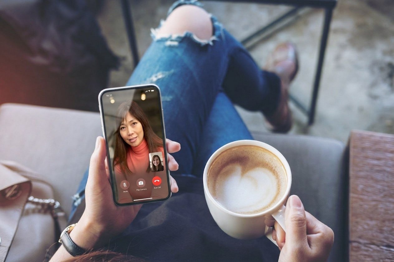 Person sitting in a cafe holding a smartphone in one hand and a cup of coffee in the other, engaged in a video call with a woman.