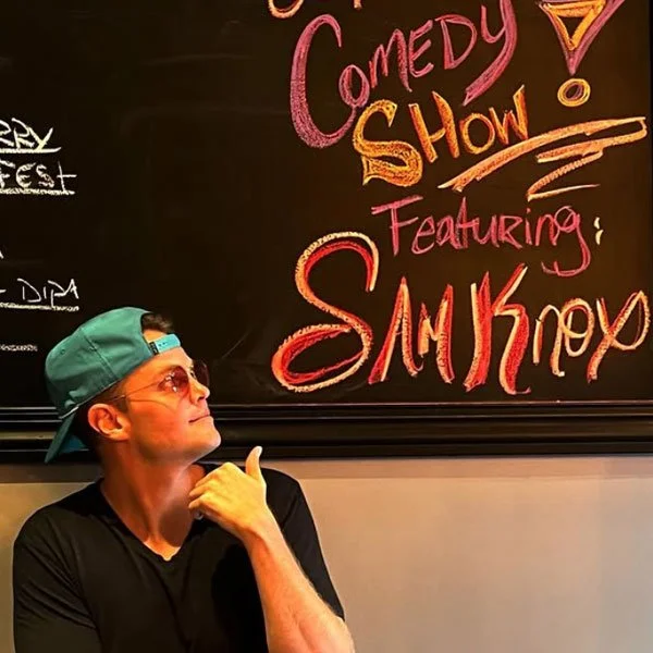 Sam Knox wearing a green cap and sunglasses, sitting with their hand on their chin, looking up at a blackboard with colorful writing about a comedy show featuring a performer named Sam K.