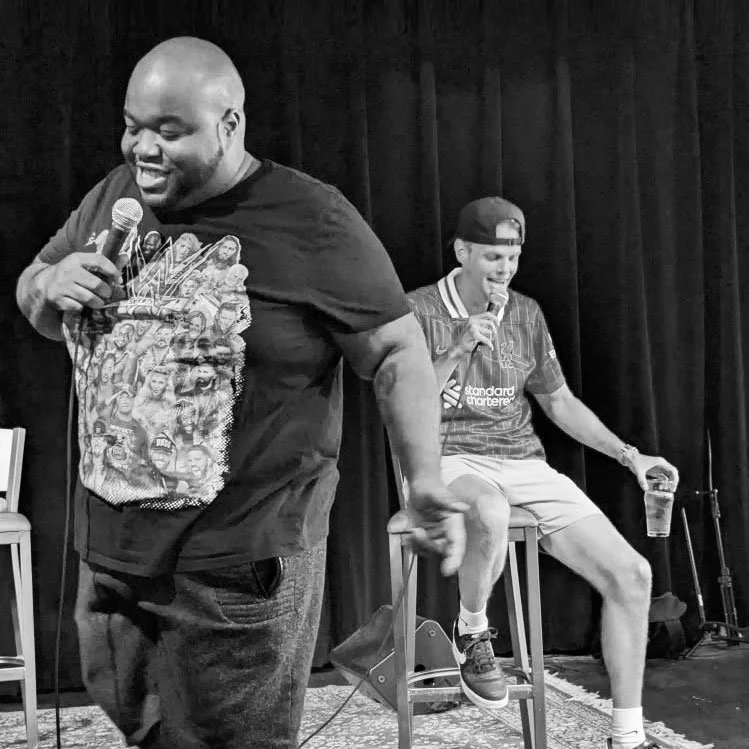 Chavez Massey and Sam Knox with microphones on a stage, one standing and one sitting, smiling during a comedy or stand-up performance in front of a dark curtain.