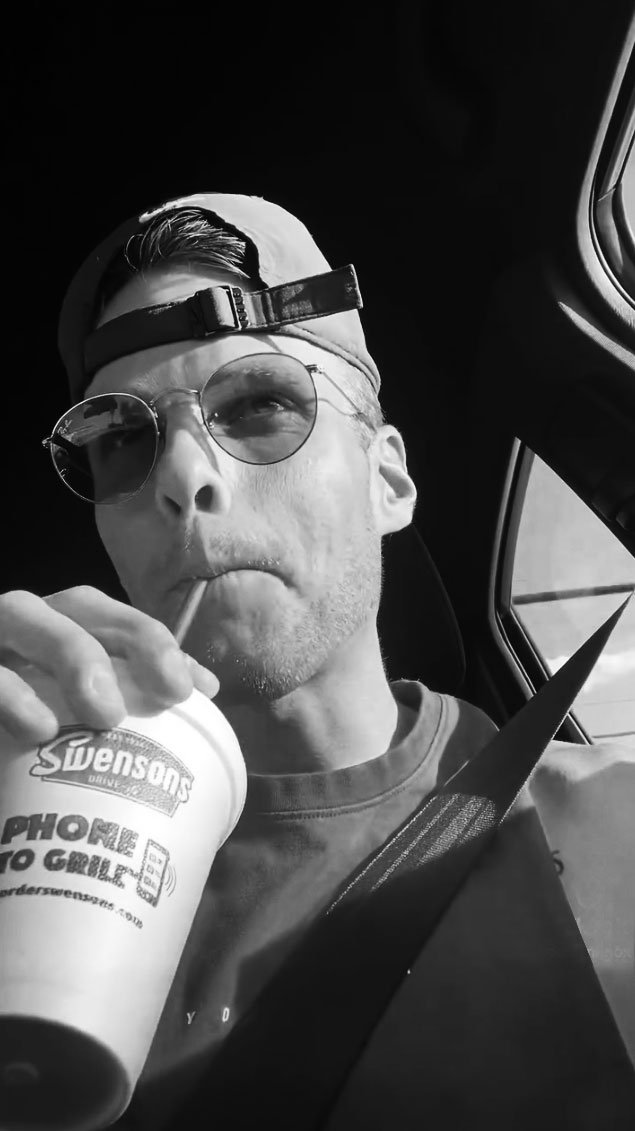 Sam Knox wearing sunglasses and a backwards baseball cap, sitting in a car, drinking from a cup with a straw.