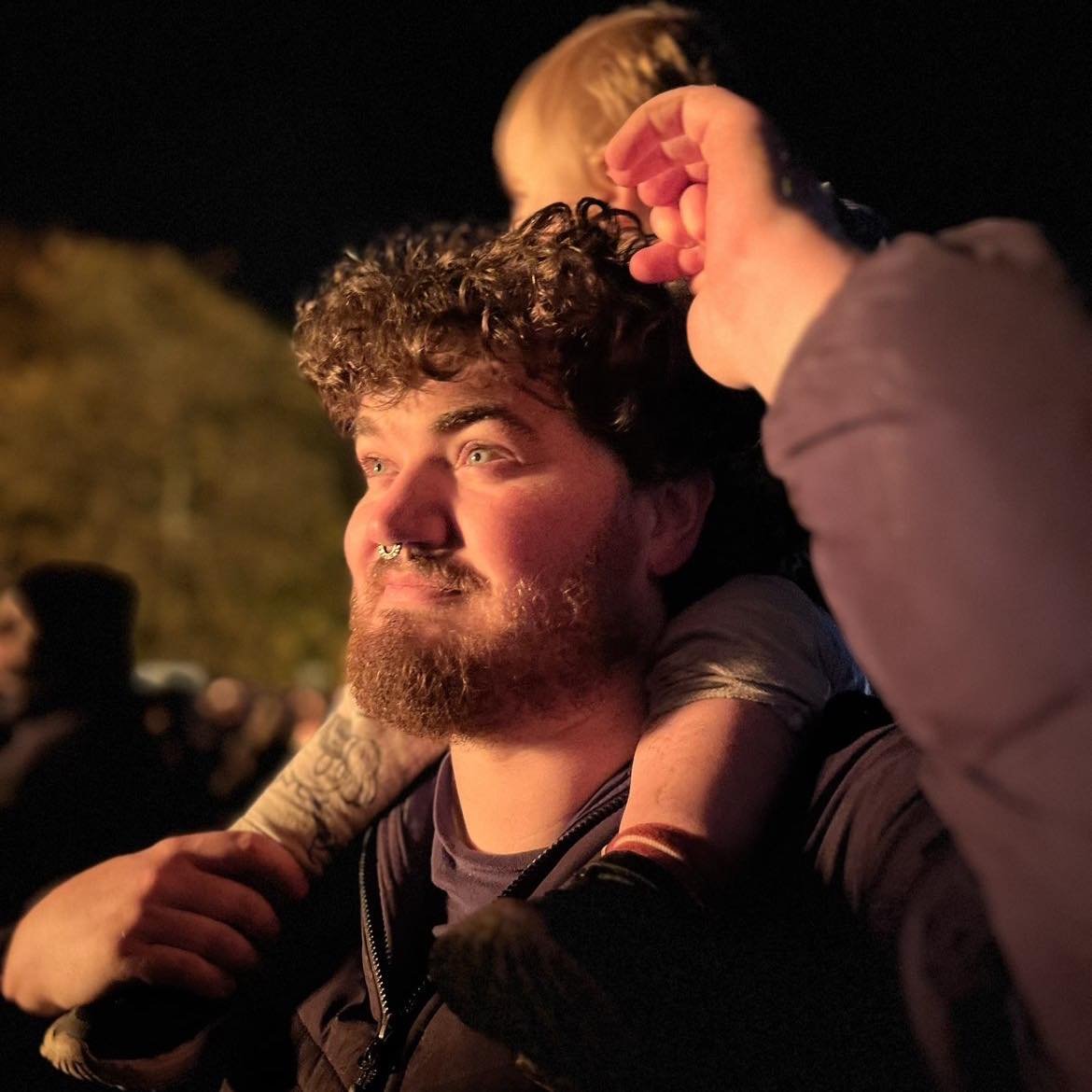 A man with curly hair and a beard holding a child on his shoulders during a nighttime outdoor event.