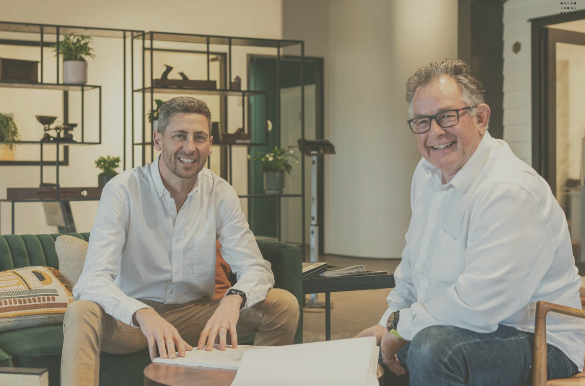 Tim and Warren sitting on a green couch and smiling in the SoundDown office with shelving and decorative plants in the background.